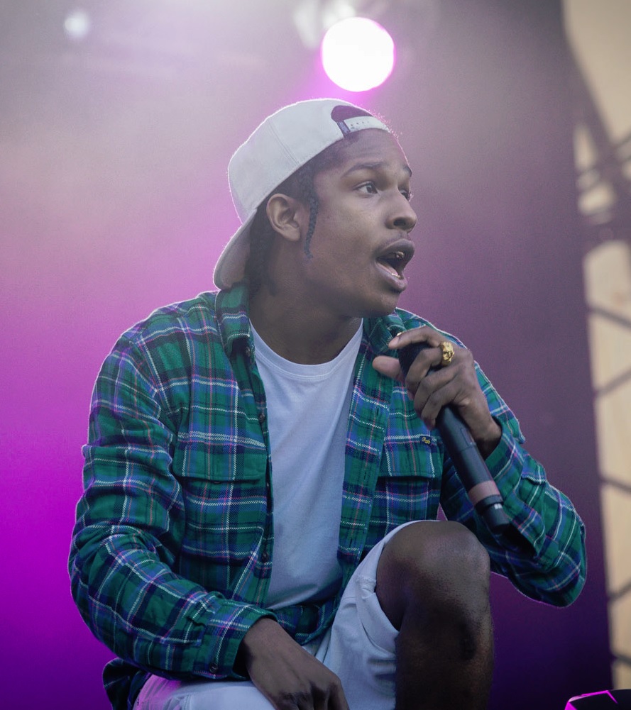 A rapper in a green plaid shirt and a backwards white cap crouches with a microphone in hand under pink stage light