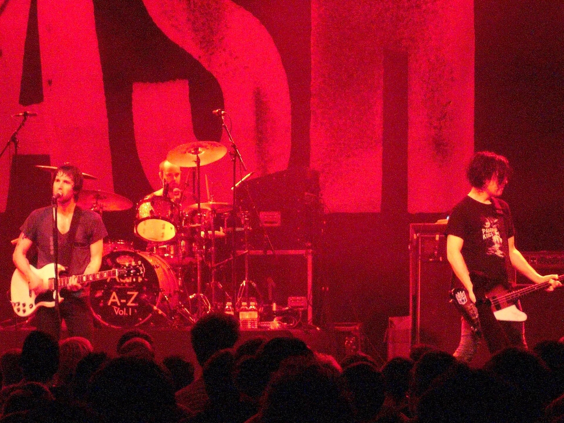 The three members of Ash perform under red stage light: a guitarist and singer on the left, a drummer behind a kit labeled A-Z Vol. 1, and a bassist on the right, with a giant glowing ASH logo on the backdrop.