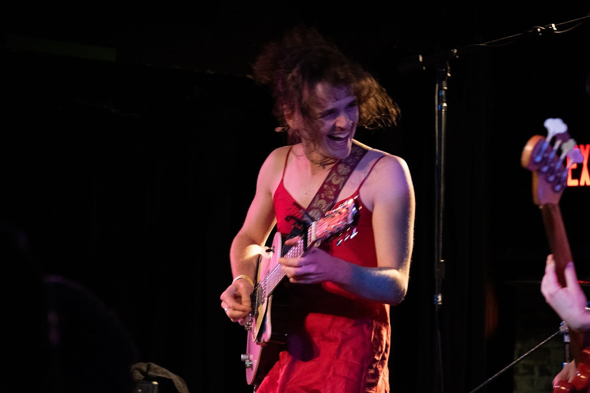 Asher White, wearing a red dress, plays a pink electric guitar and sings on stage with her hair in motion.