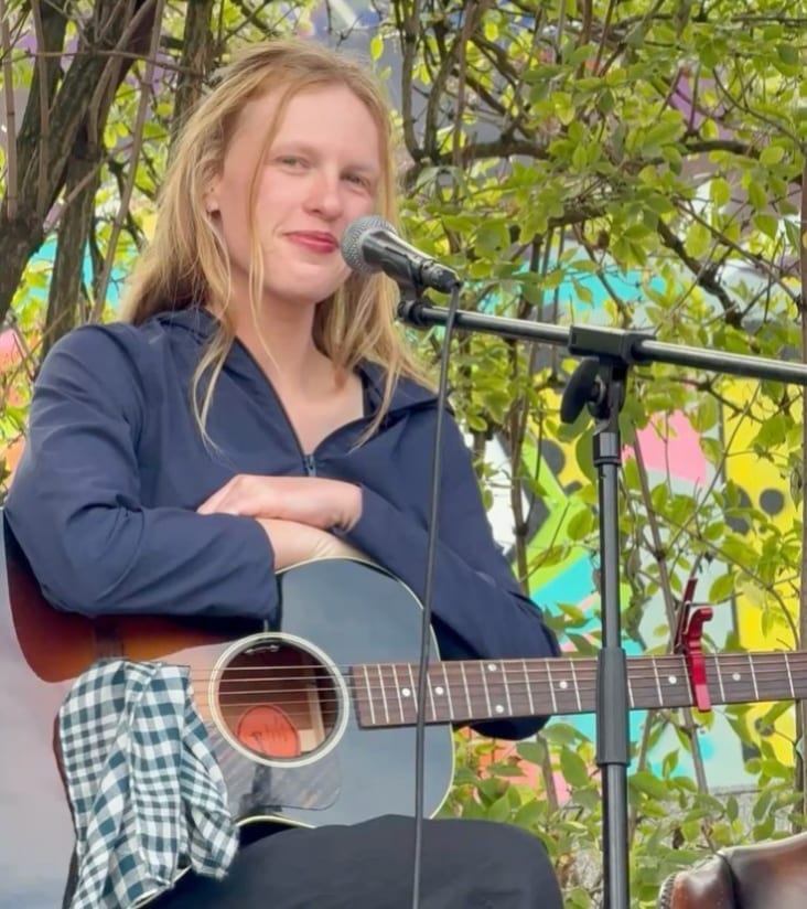 Audrey Hobert smiling while sitting outdoors with an acoustic guitar in front of a microphone stand, with green foliage and a colorful mural behind her.