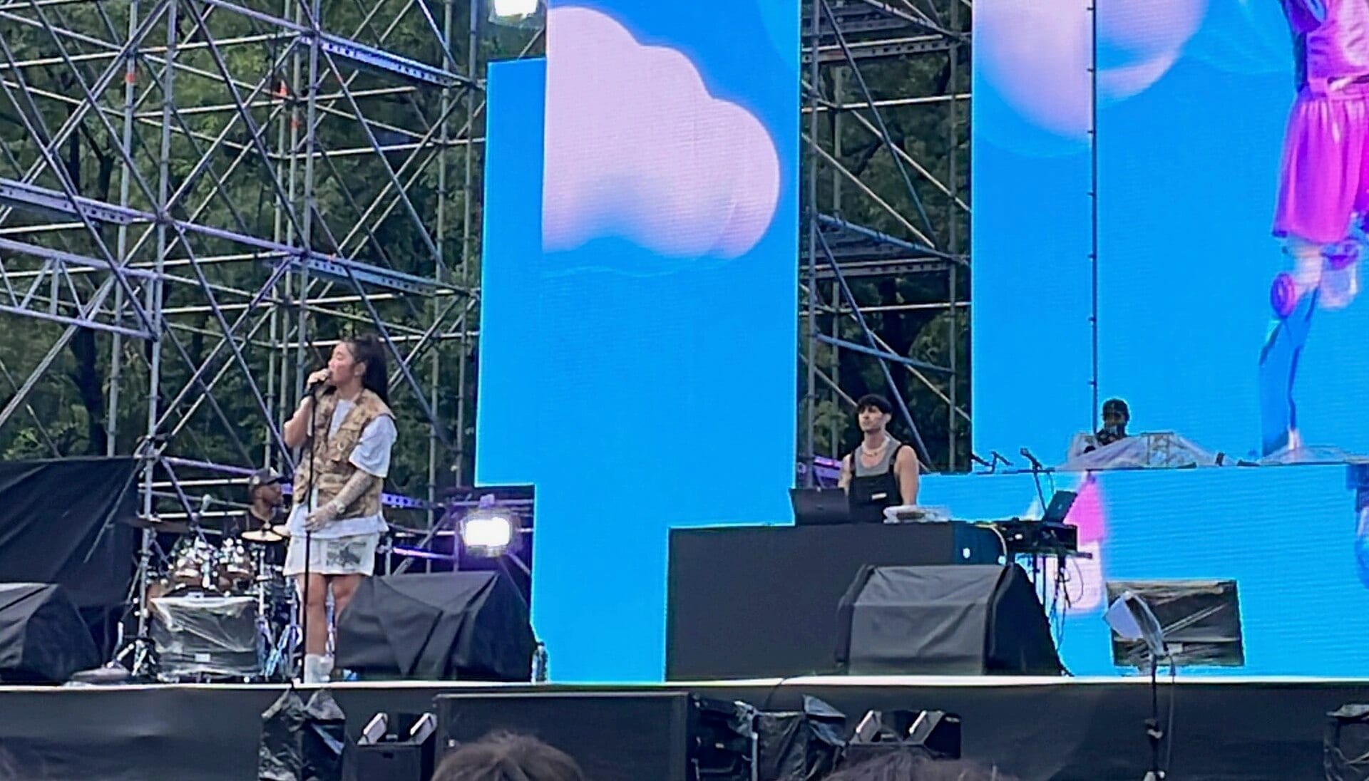 Audrey Nuna sings into a microphone on an outdoor festival stage, wearing a patterned vest over a white shirt, with a drummer and screens showing blue sky visuals behind her.