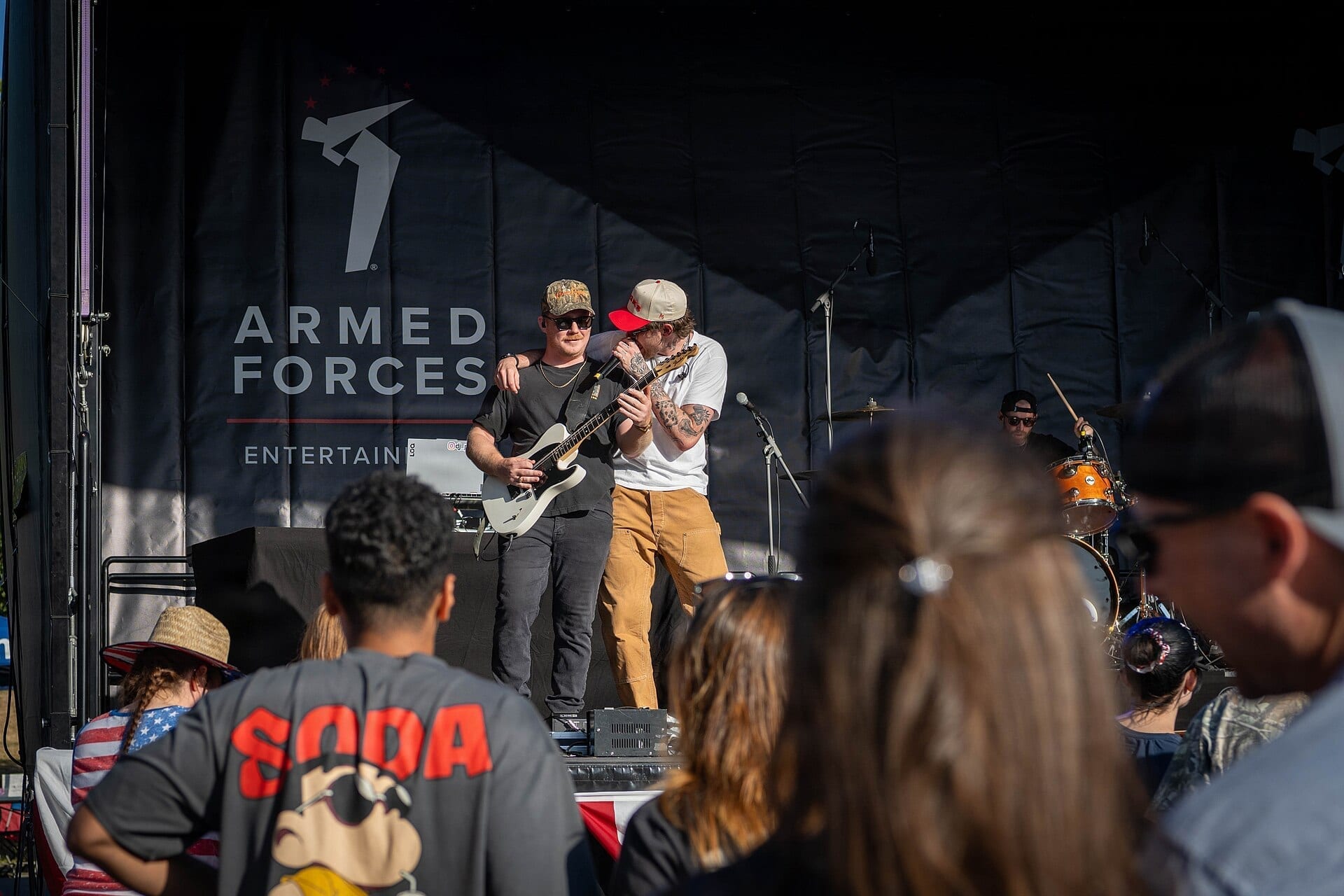 Austin Snell sings into a microphone on an outdoor stage next to a guitarist, in front of an Armed Forces Entertainment backdrop, with a crowd watching below.
