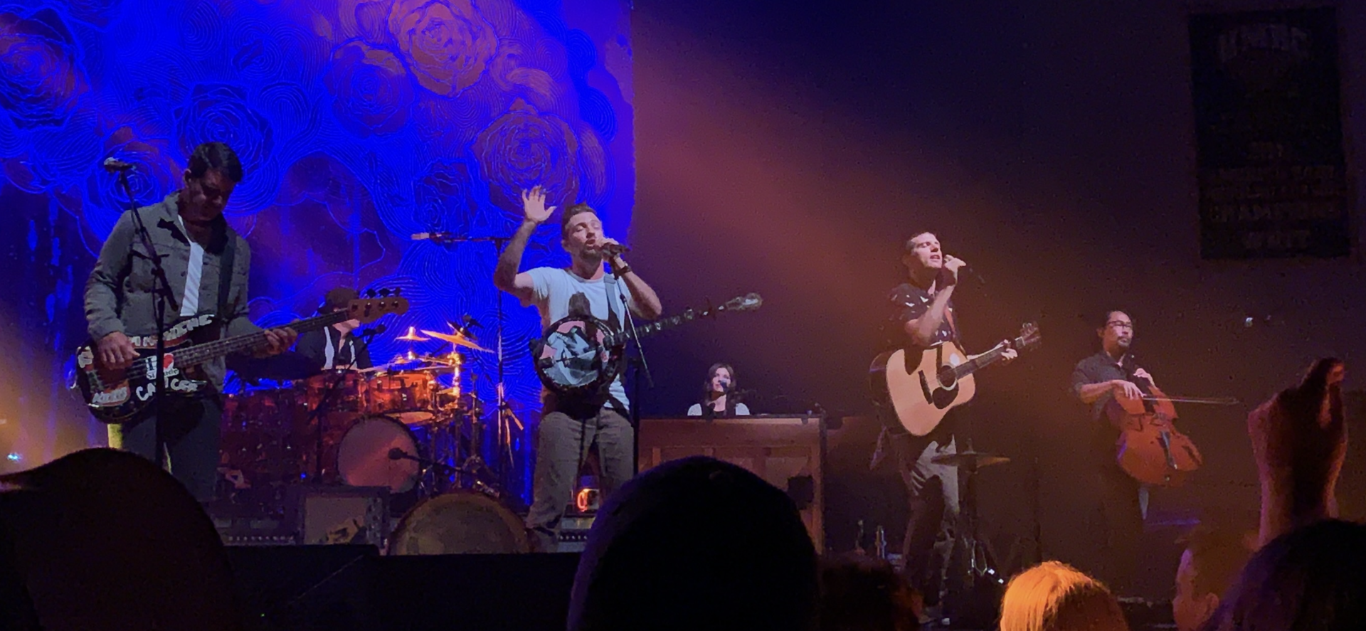The Avett Brothers spread across a wide stage: a bassist at left, a banjo player singing with one hand raised in the centre, an acoustic guitarist and a cellist at right.