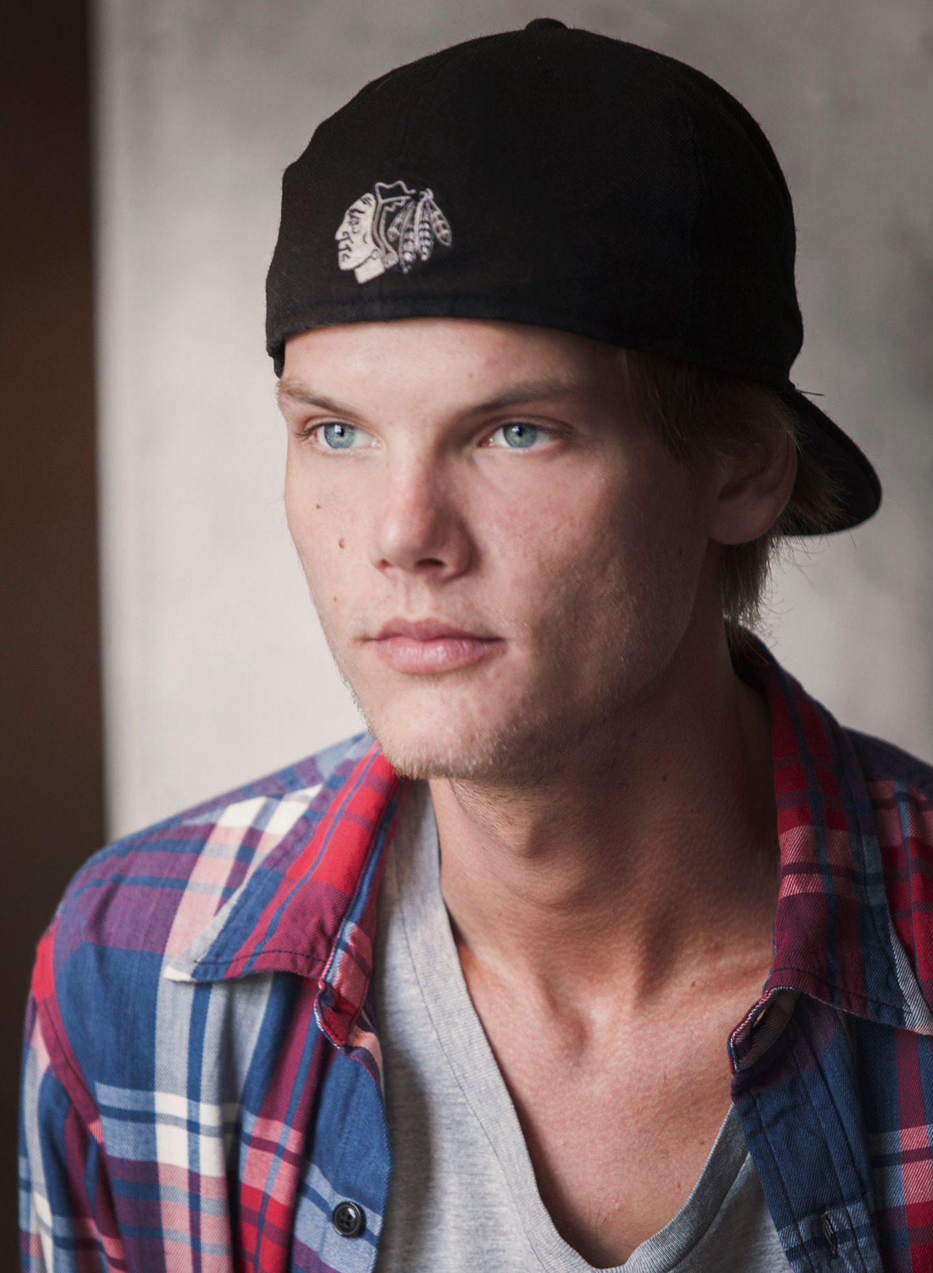 Avicii sits at a table in a baseball cap and a checked flannel shirt over a white tee, looking to one side.