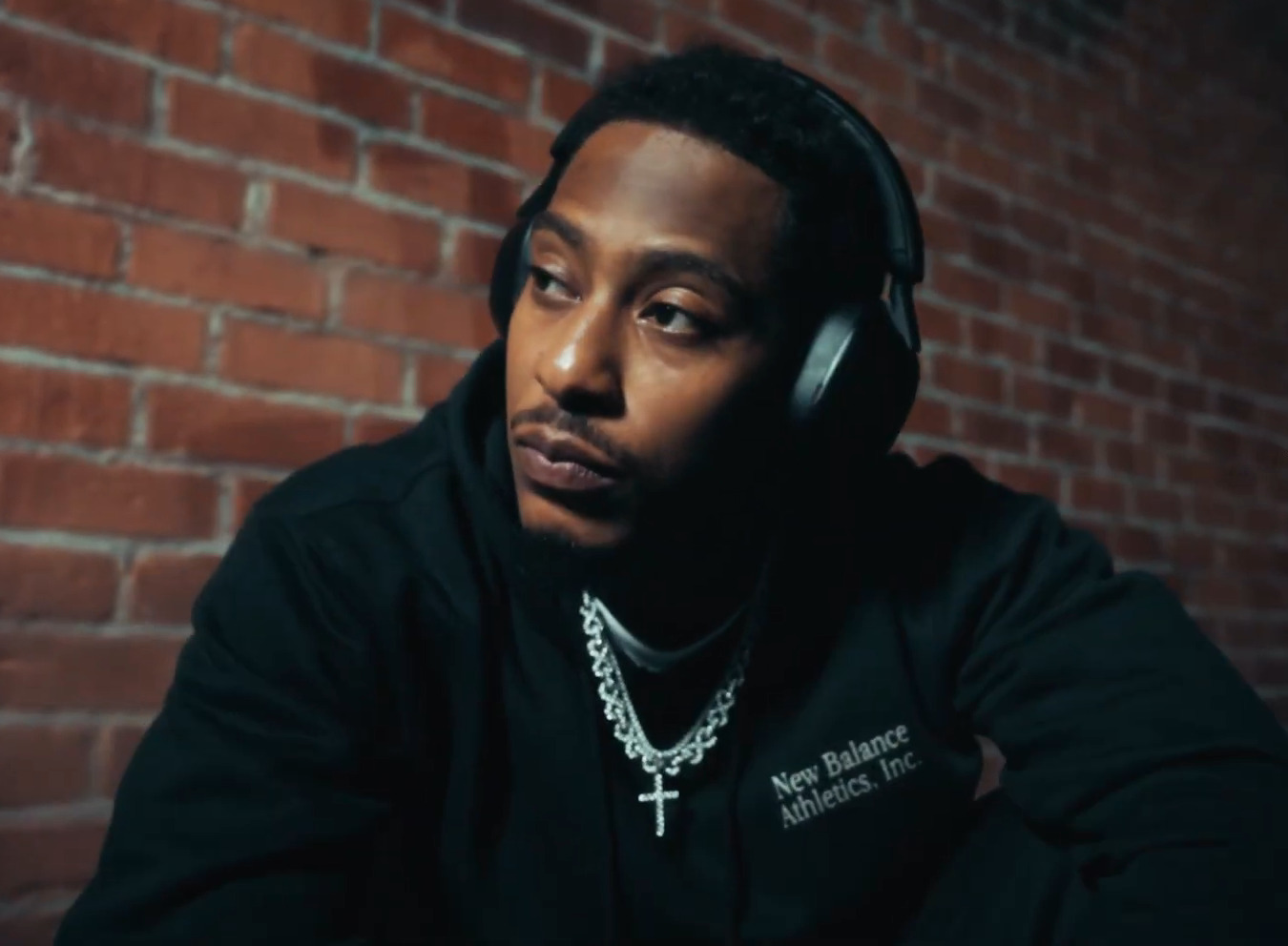 AZ Chike stands against a brick wall in a grey hooded top and over-ear headphones, looking to one side.