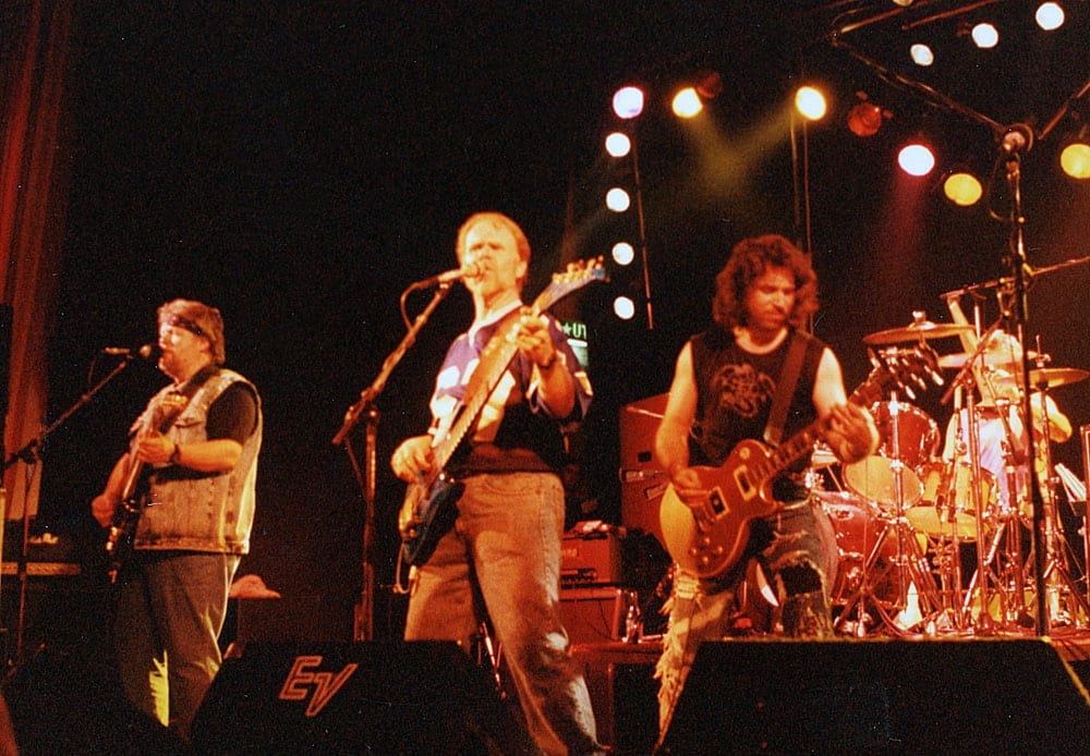 Four musicians play guitars and drums on a small stage under warm lights, a monitor wedge marked EV at the front.