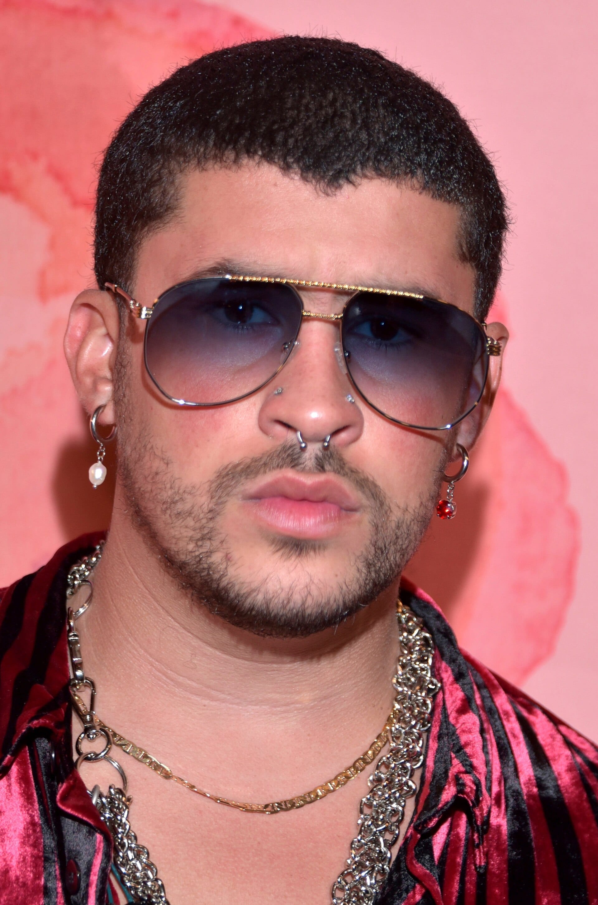 A close portrait of Bad Bunny in tinted aviator sunglasses, a septum ring and drop earrings, wearing a red and black striped velvet shirt and layered chains.
