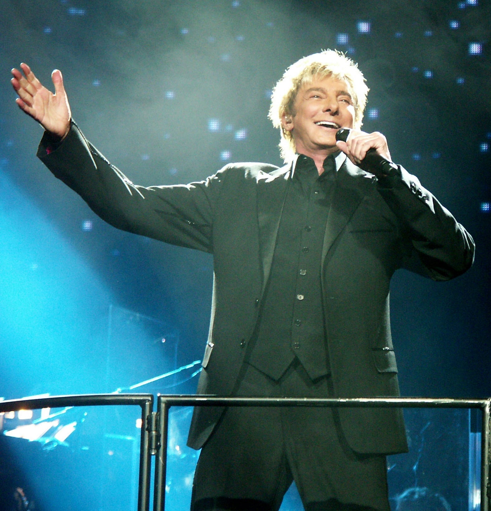 Barry Manilow sings into a handheld microphone on a blue-lit arena stage, one arm thrown wide, wearing a black three-piece suit and open-collar shirt.