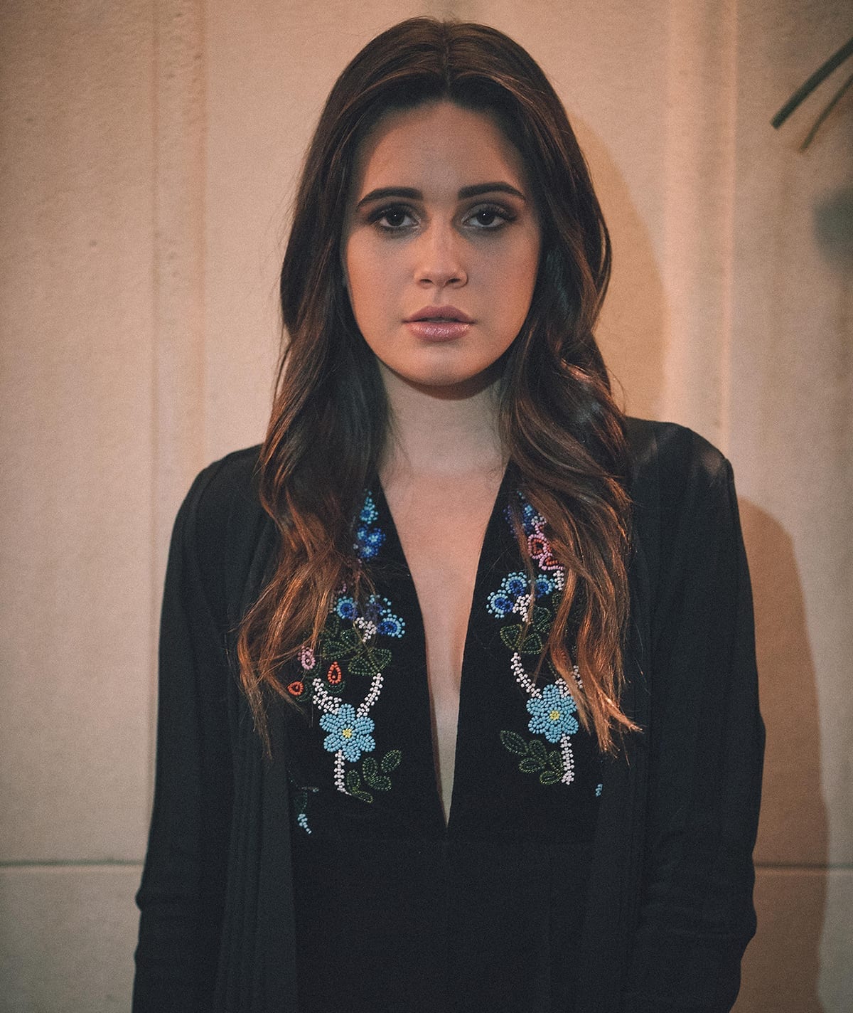 Bea Miller stares directly at the camera in a close portrait, wearing a black top with beaded floral embroidery, her long dark hair loose over her shoulders.