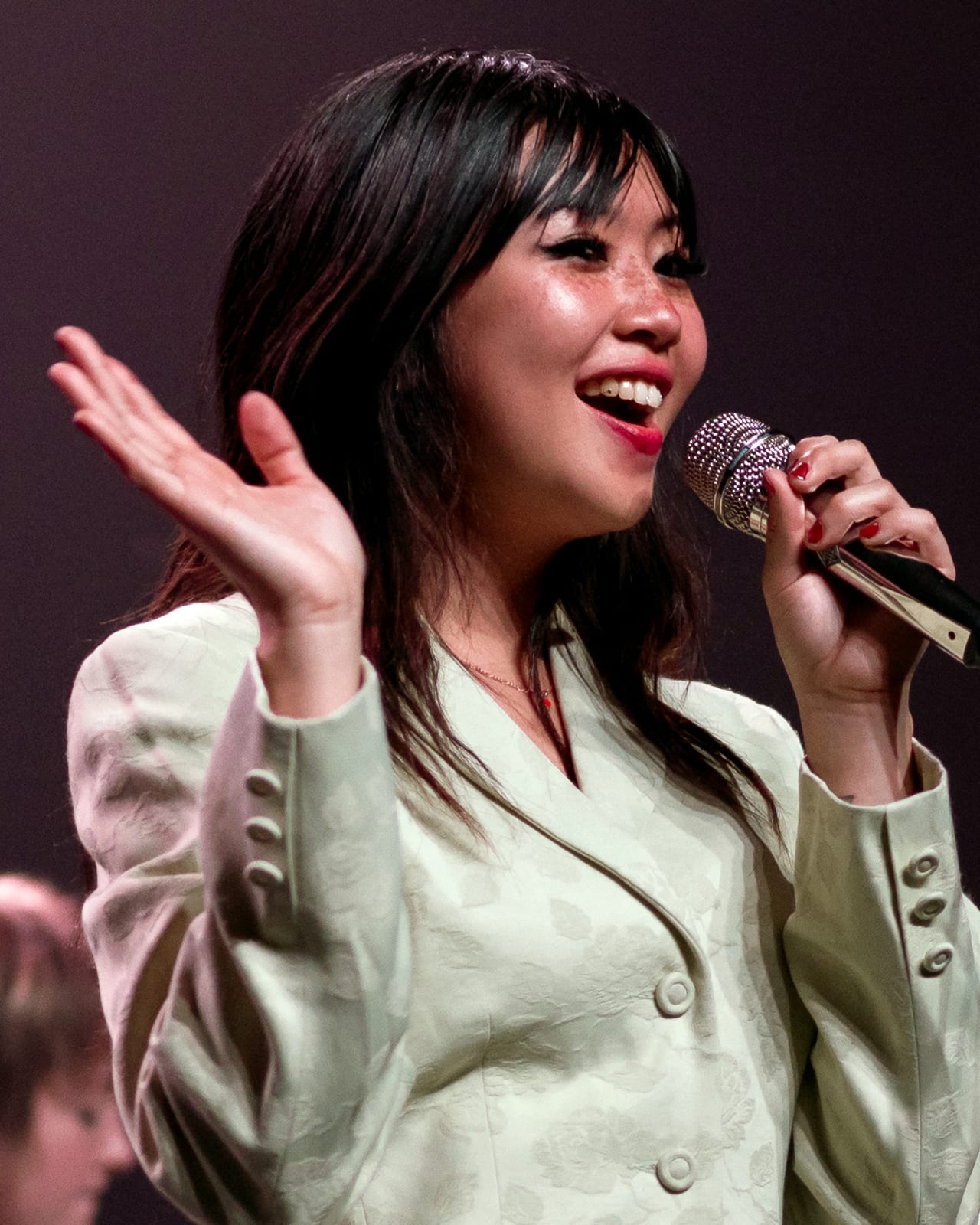 Beabadoobee sings into a handheld microphone with one hand raised, in a pale green jacket, her dark hair cut in a fringe.
