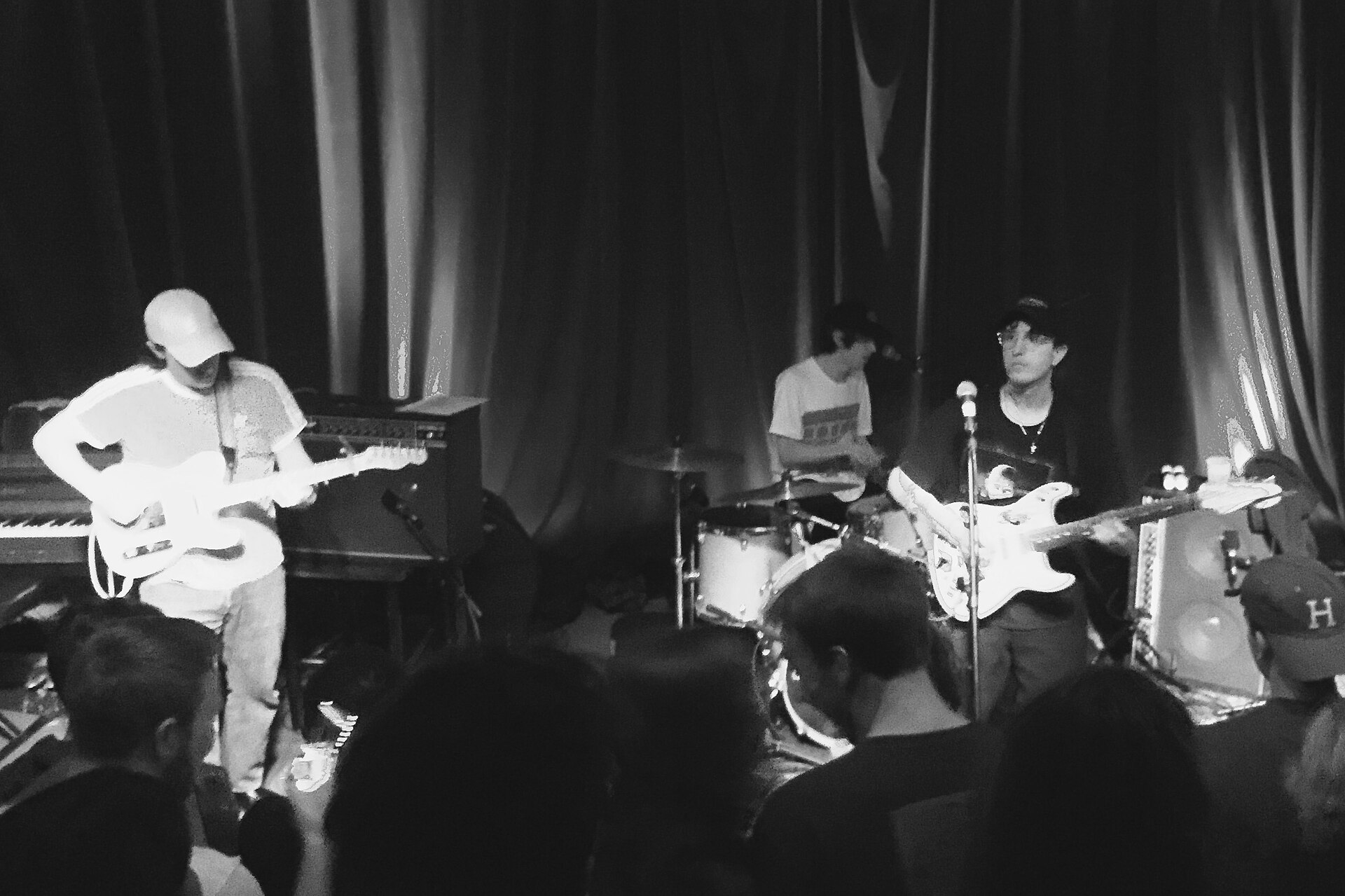Black and white shot of Beach Fossils on a small club stage, two guitarists and a drummer in front of heavy stage curtains, seen over the heads of the crowd
