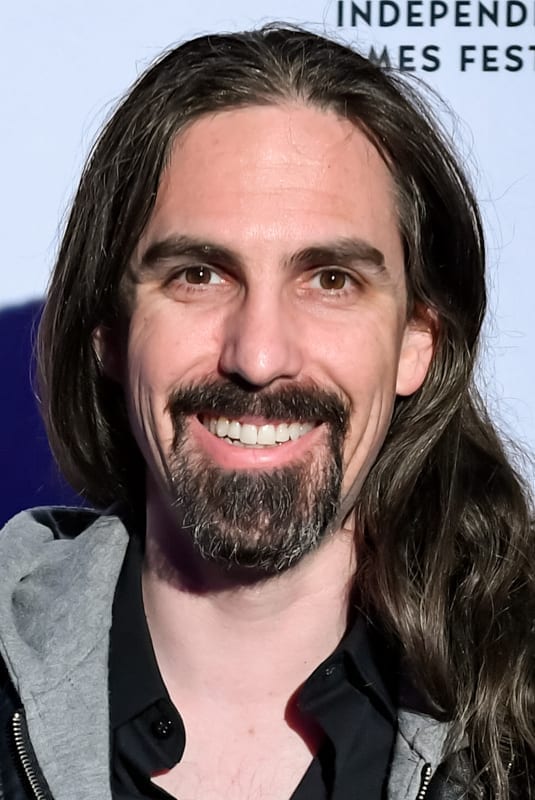 Bear McCreary, with long dark hair and a beard, smiles at a step-and-repeat event.