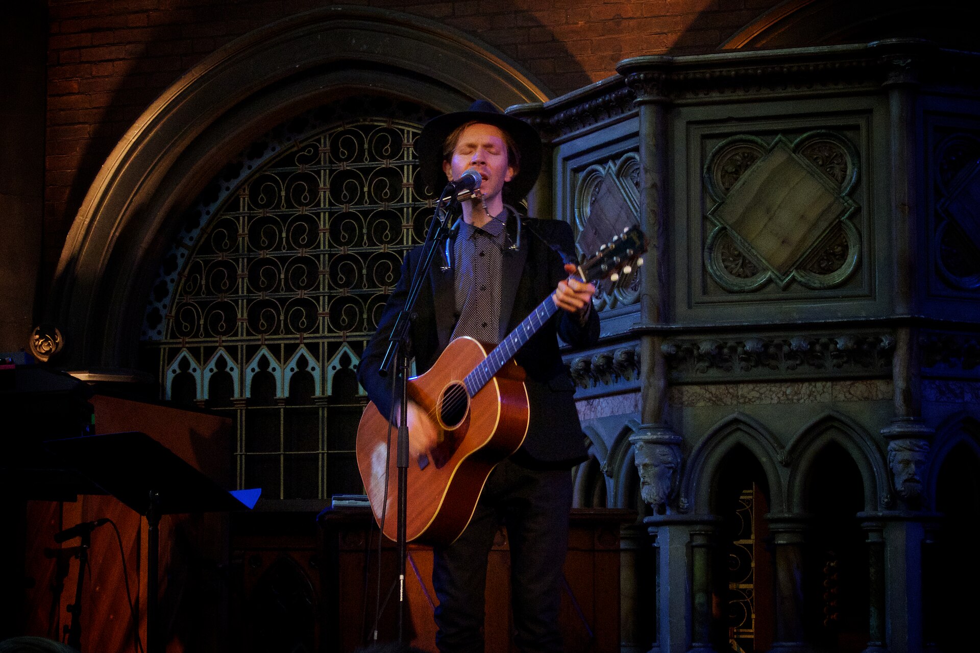 Beck sings into a microphone with his eyes closed, playing an acoustic guitar in a black hat and jacket in front of a Gothic stone pulpit and arched window.