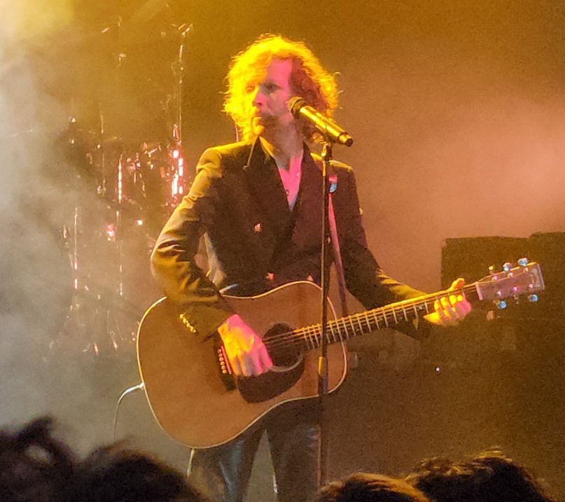 A curly-haired singer in a dark jacket plays an acoustic guitar at a microphone stand, haze and warm stage light around him