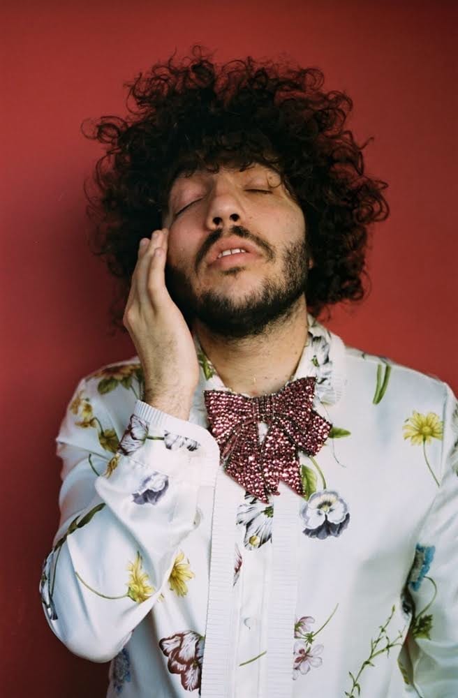 Benny Blanco rests a hand against his cheek with his eyes closed, wearing a floral shirt and a sequinned bow tie against a red background.