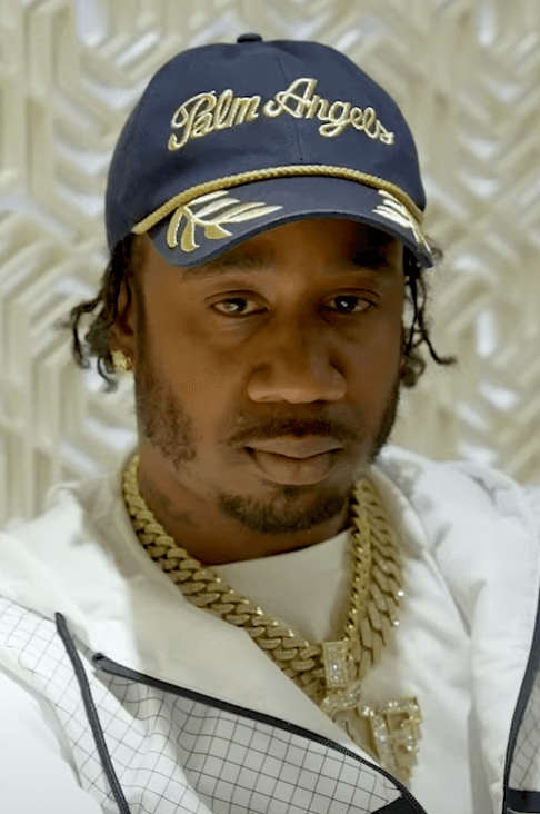 Benny The Butcher poses in close-up wearing a navy Palm Angels cap, layered gold chains including a diamond-encrusted pendant, and a white jacket.