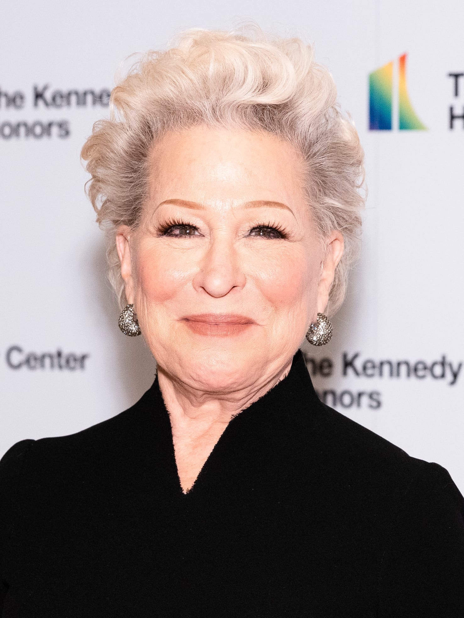 Bette Midler, with short platinum hair and sparkling drop earrings, smiles in a black high-collared outfit in front of a Kennedy Center Honors backdrop.
