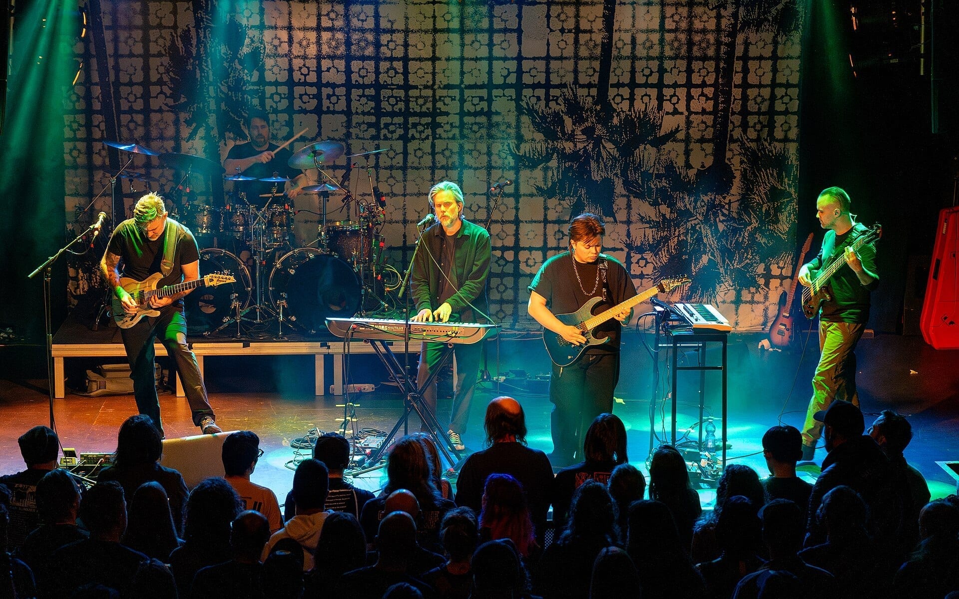 The five members of Between the Buried and Me perform on a green-lit stage with guitars, a keyboard and drums, in front of a crowd.