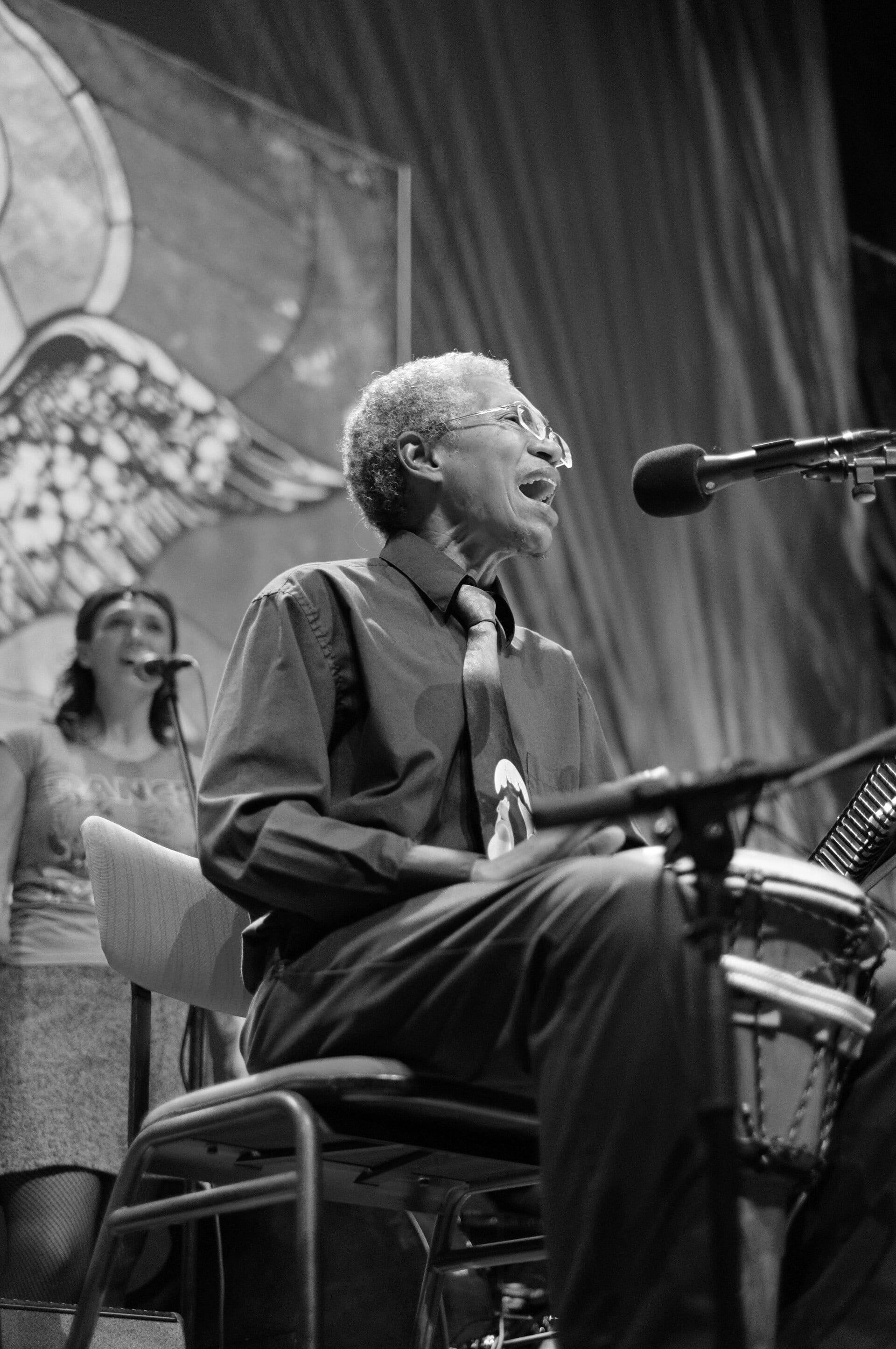 A black-and-white photo of Beverly Glenn-Copeland singing joyfully into a microphone while seated on stage, wearing glasses and a tie.