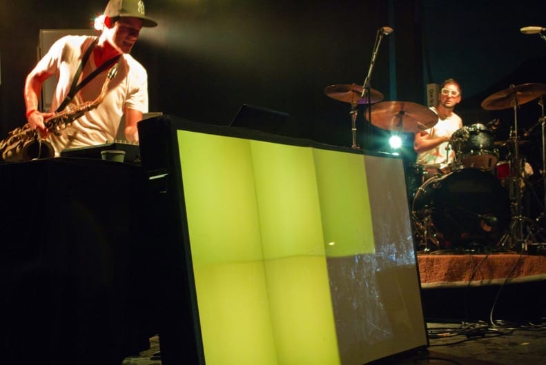 Big Gigantic perform on stage, with one member playing saxophone and the other playing drums behind him.