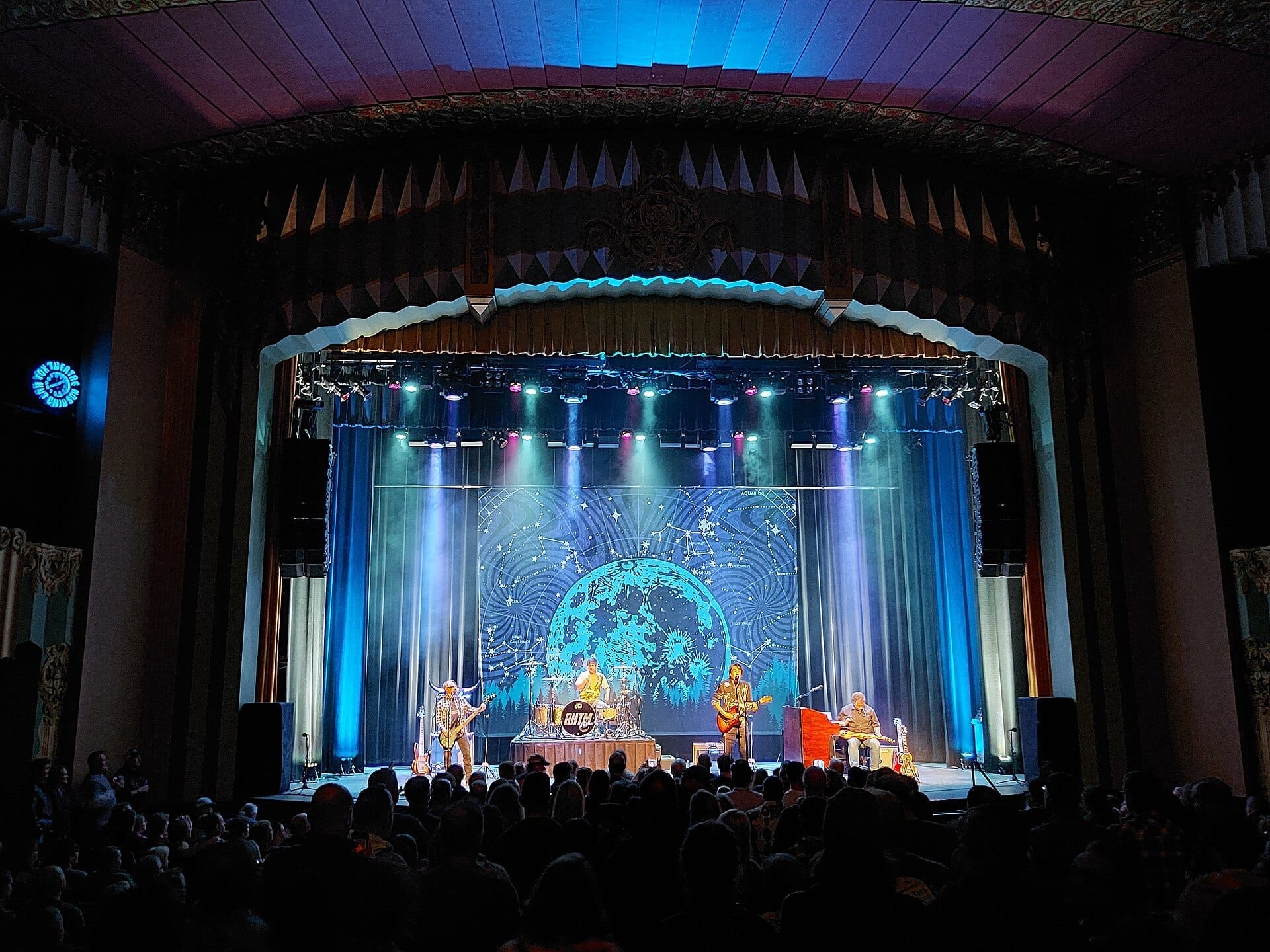 Big Head Todd and the Monsters perform on a theater stage lit in blue with a starry moon backdrop, seen from the audience beneath an ornate balcony.
