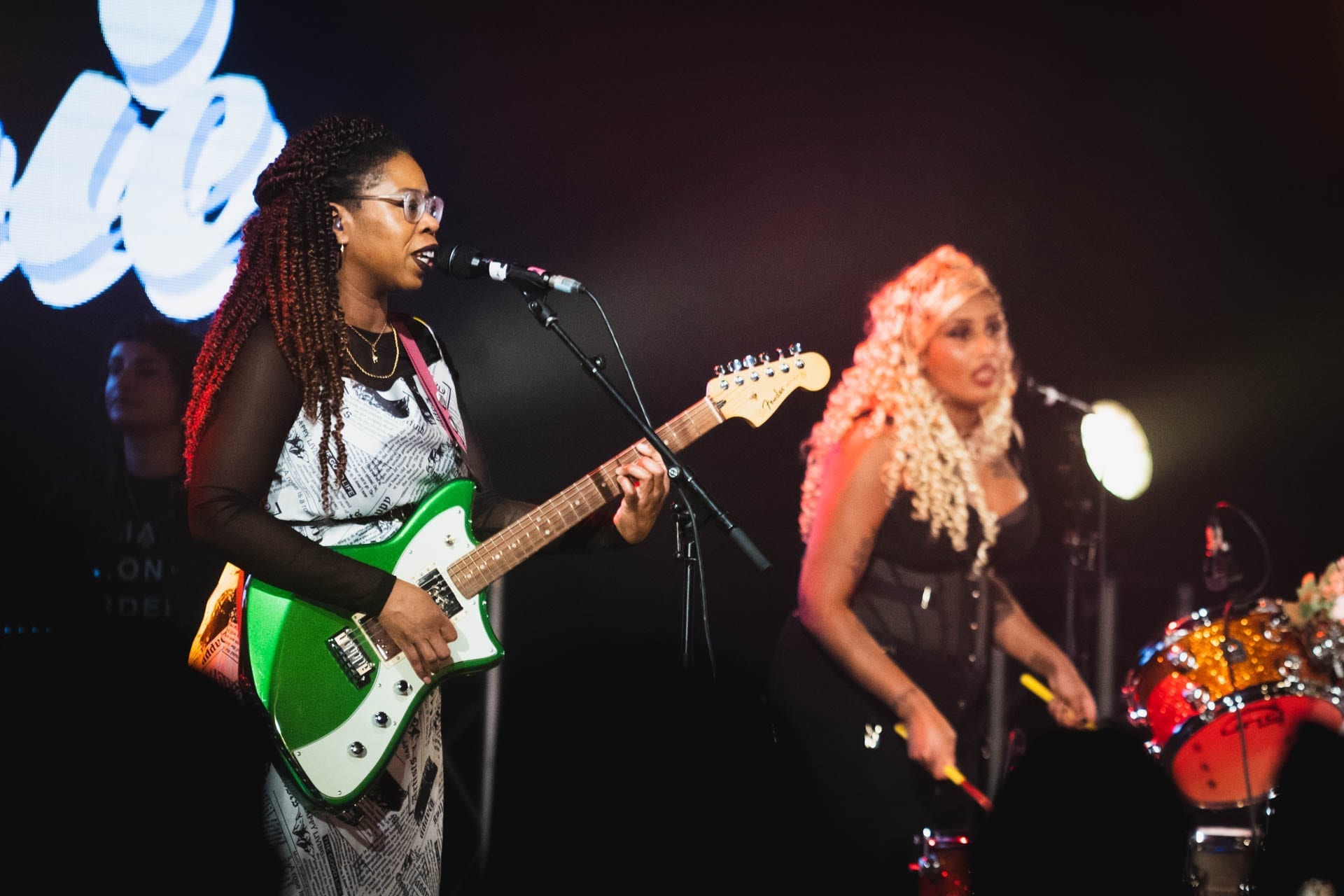Big Joanie's guitarist singing into a microphone and playing a green electric guitar on stage, with a bassist and drummer performing behind her.