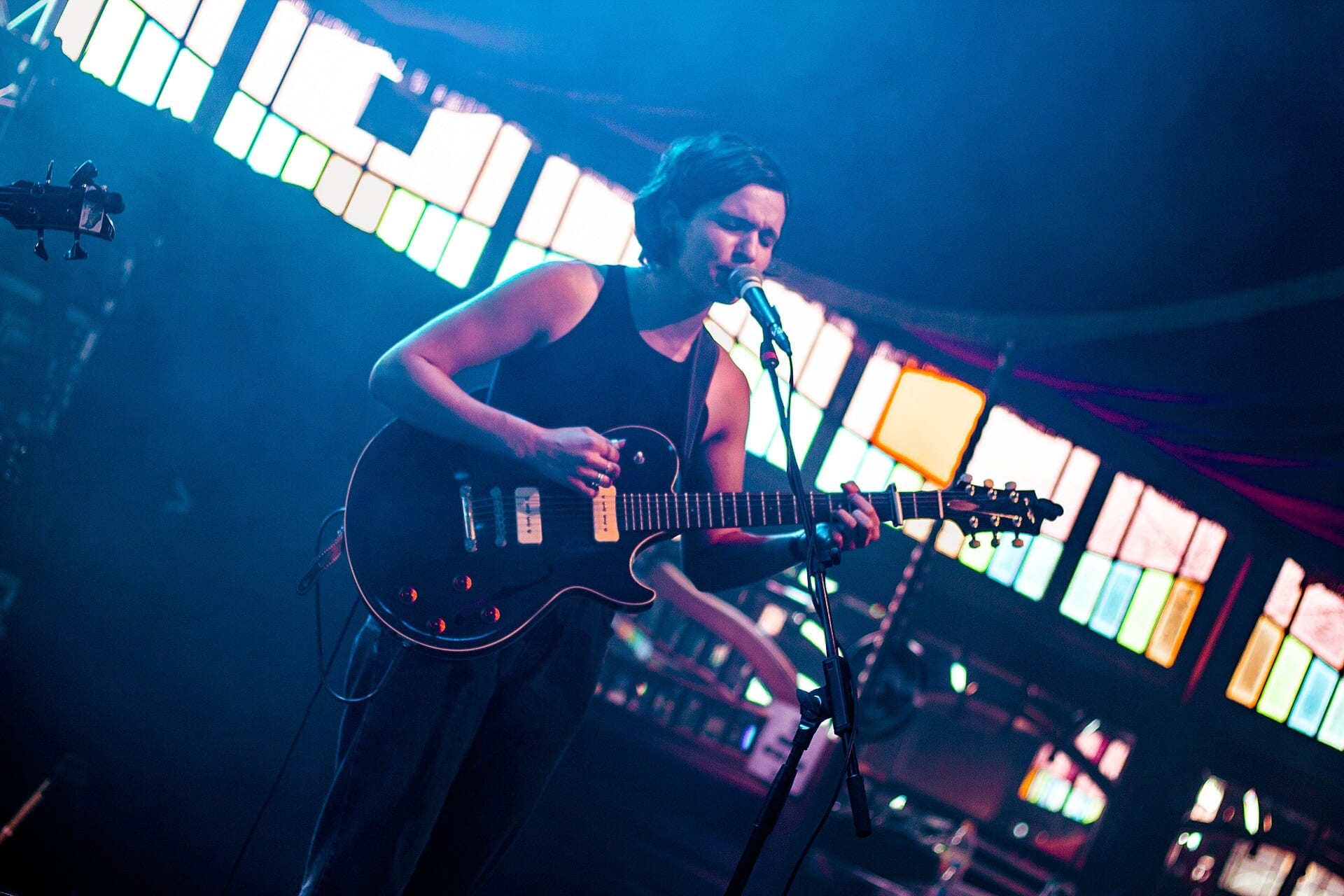 A singer leans into a microphone while playing a black electric guitar, lit blue against a wall of white stage panels.