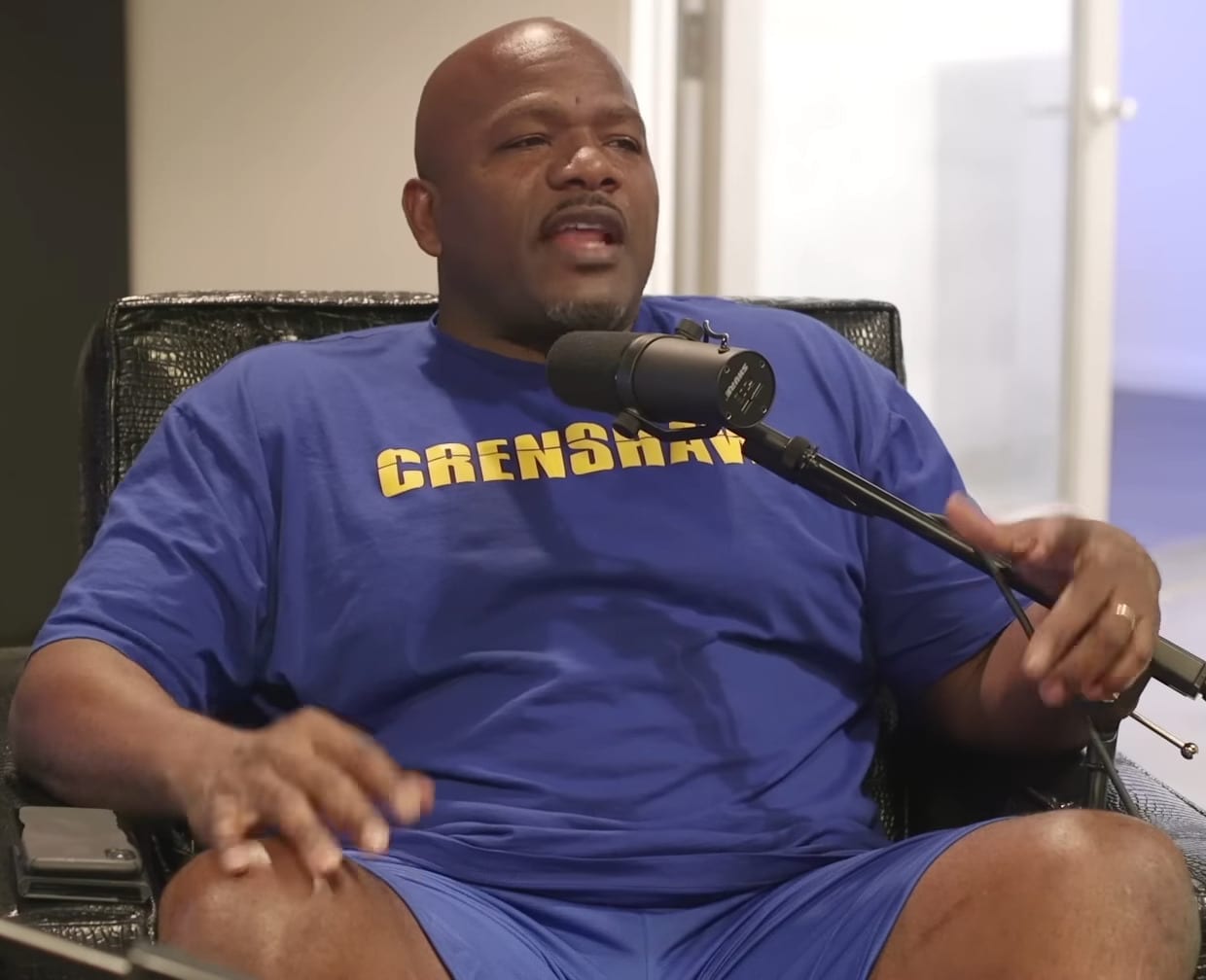 Big U sits in a chair speaking into a podcast microphone, wearing a blue Crenshaw T-shirt.