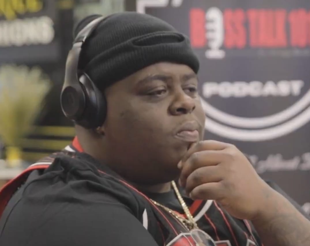 BigXthaPlug rests a hand near his chin, wearing a black beanie and studio headphones in front of a podcast backdrop.
