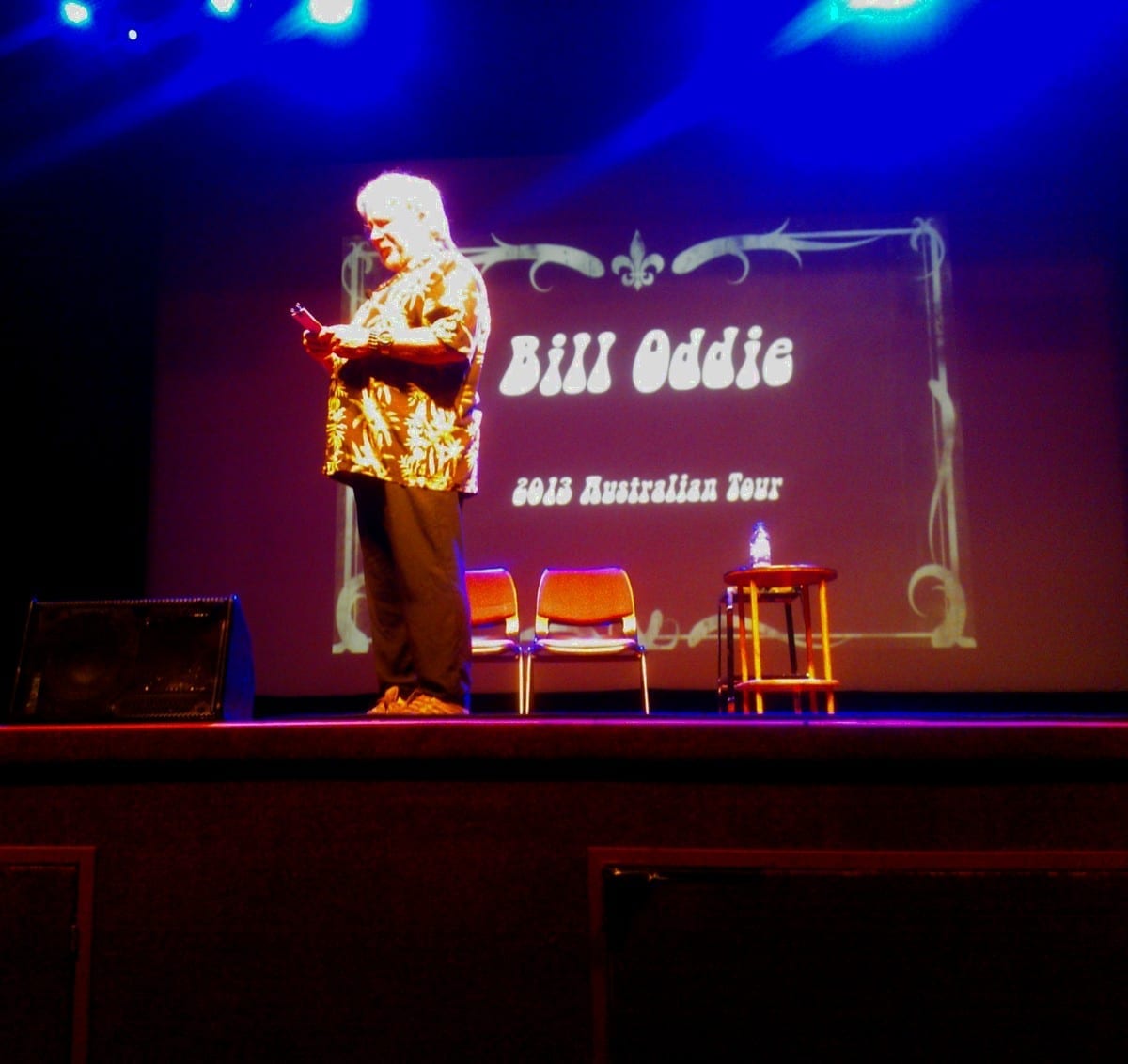 Bill Oddie stands on a stage holding a phone, lit in blue and pink stage lighting, in front of a screen reading Bill Oddie, 2013 Australian Tour.
