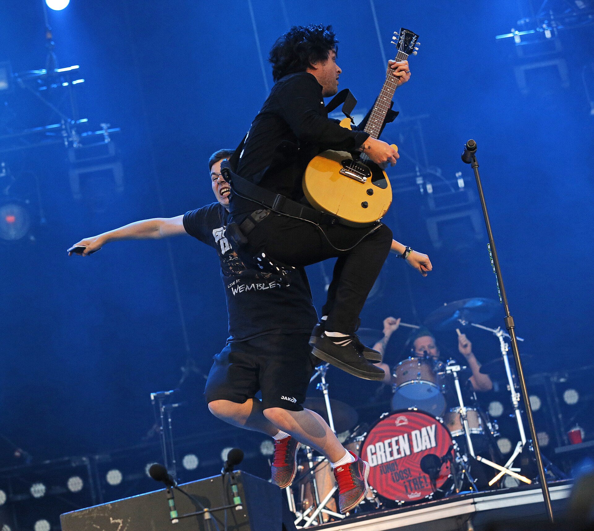 Billie Joe Armstrong leaps off the stage lip with his guitar, arms out, over a barrier crowded with fans and photographers.