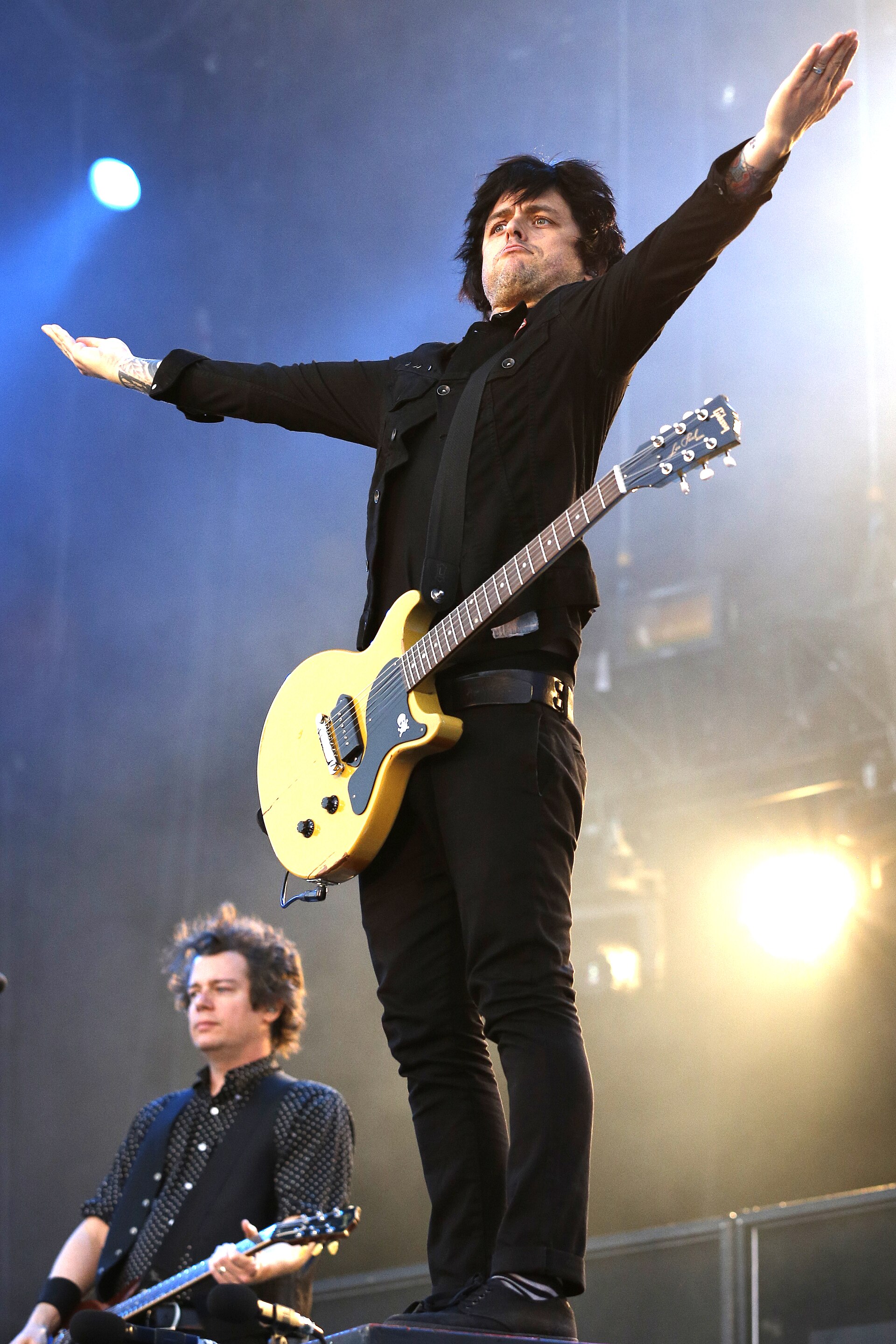 Billie Joe Armstrong stands with both arms flung wide at a microphone stand, a yellow electric guitar slung at his hip, on a festival stage.