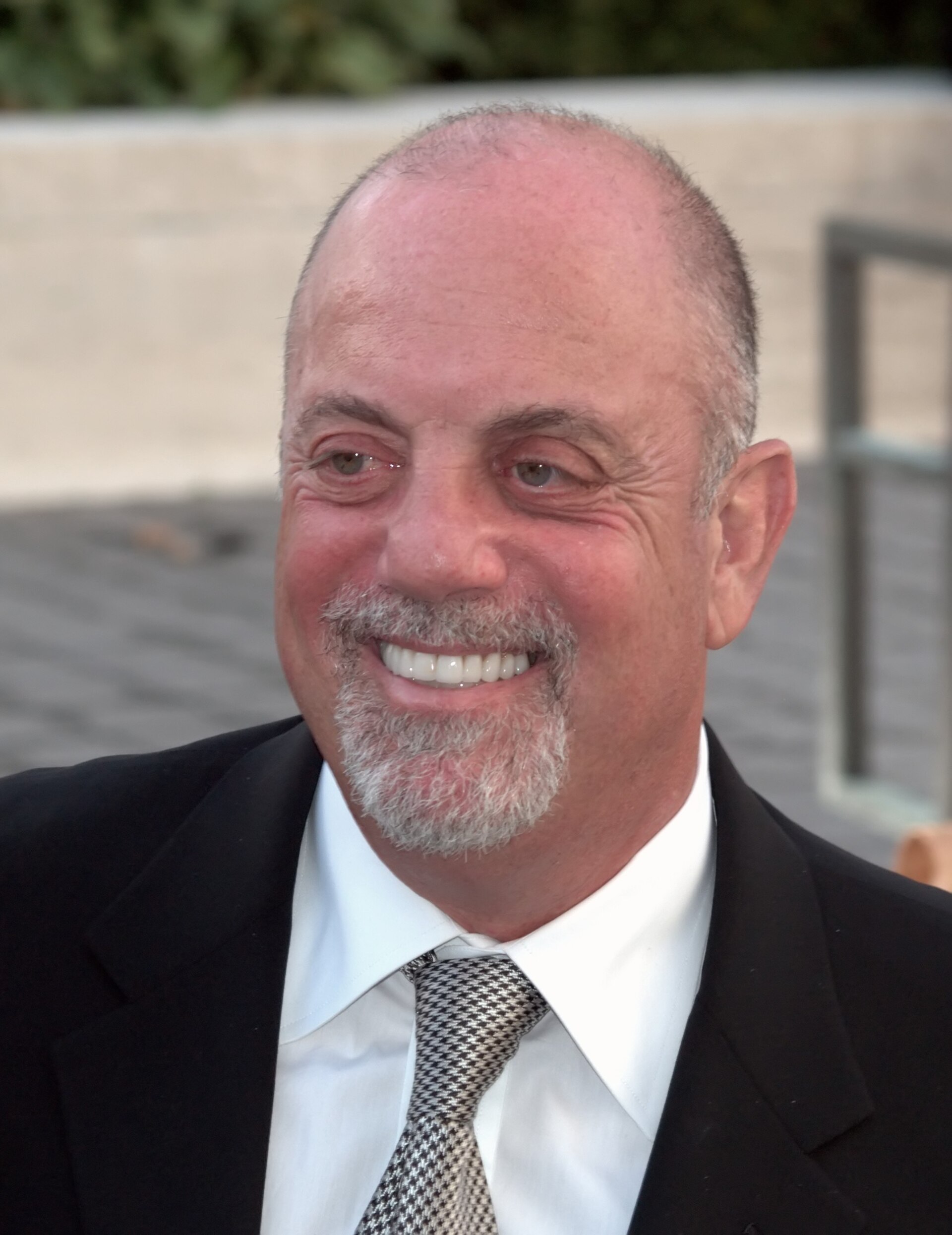 A close portrait of a smiling Billy Joel outdoors in a dark suit, white shirt and houndstooth tie, with a grey goatee and closely cropped hair.