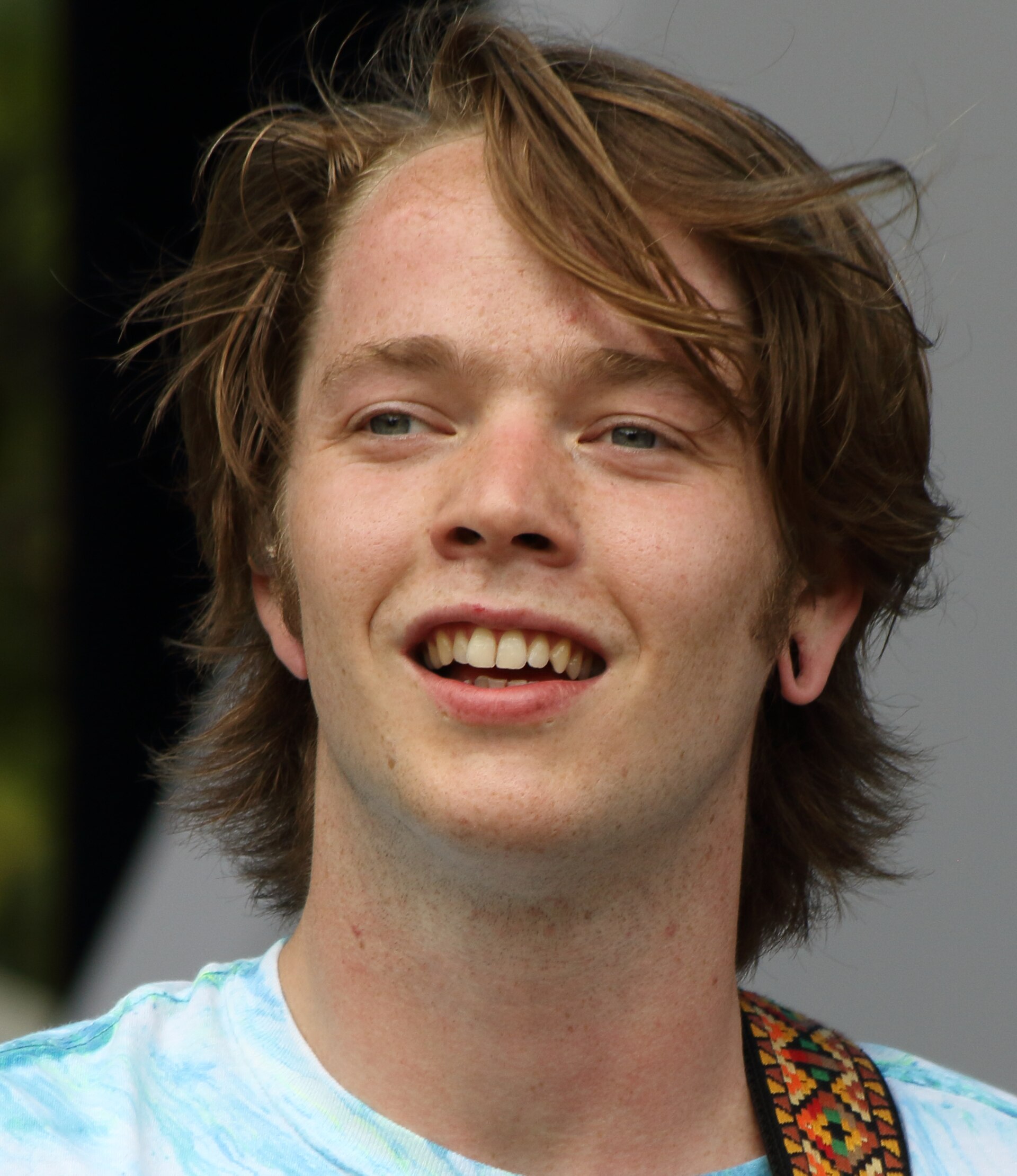 Close portrait of Billy Strings smiling, shaggy fair hair blown across his forehead, a woven guitar strap over one shoulder.