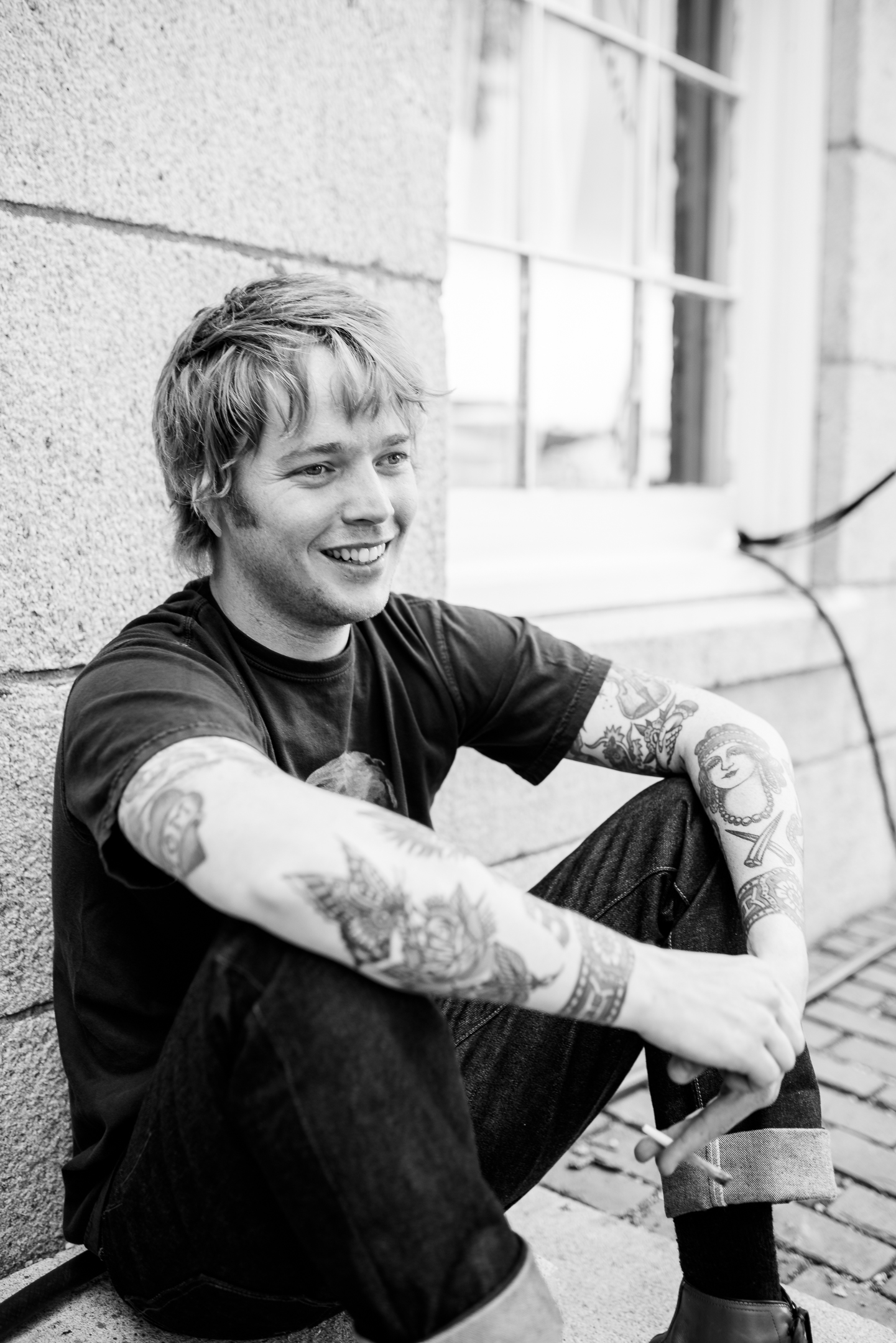 Black and white portrait of Billy Strings sitting on a step against a stone wall, knees drawn up, smiling off to one side, tattoos covering both forearms.