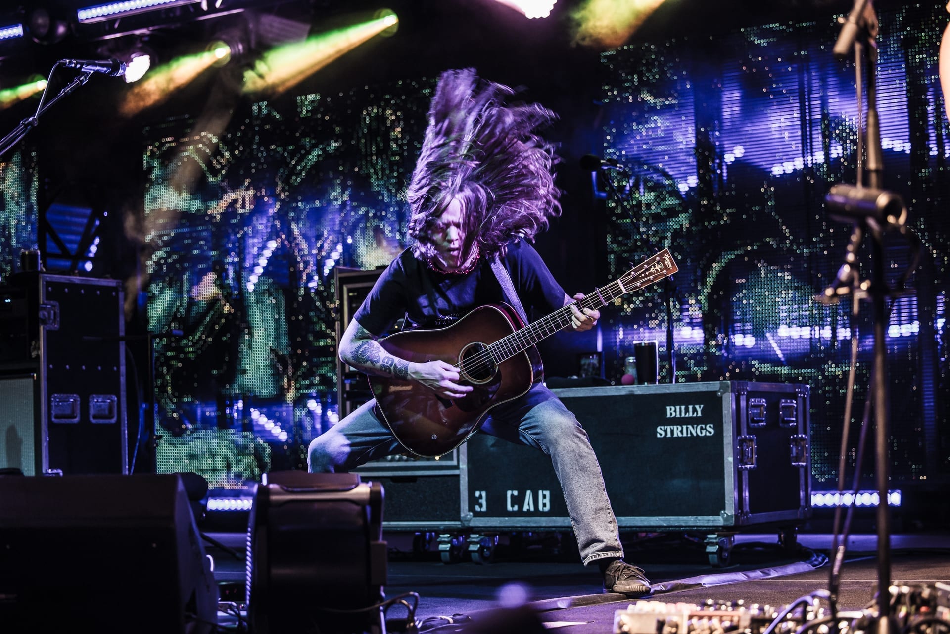 Billy Strings throws his hair back mid-song while playing an acoustic guitar, legs braced wide, a road case printed with his name behind him.