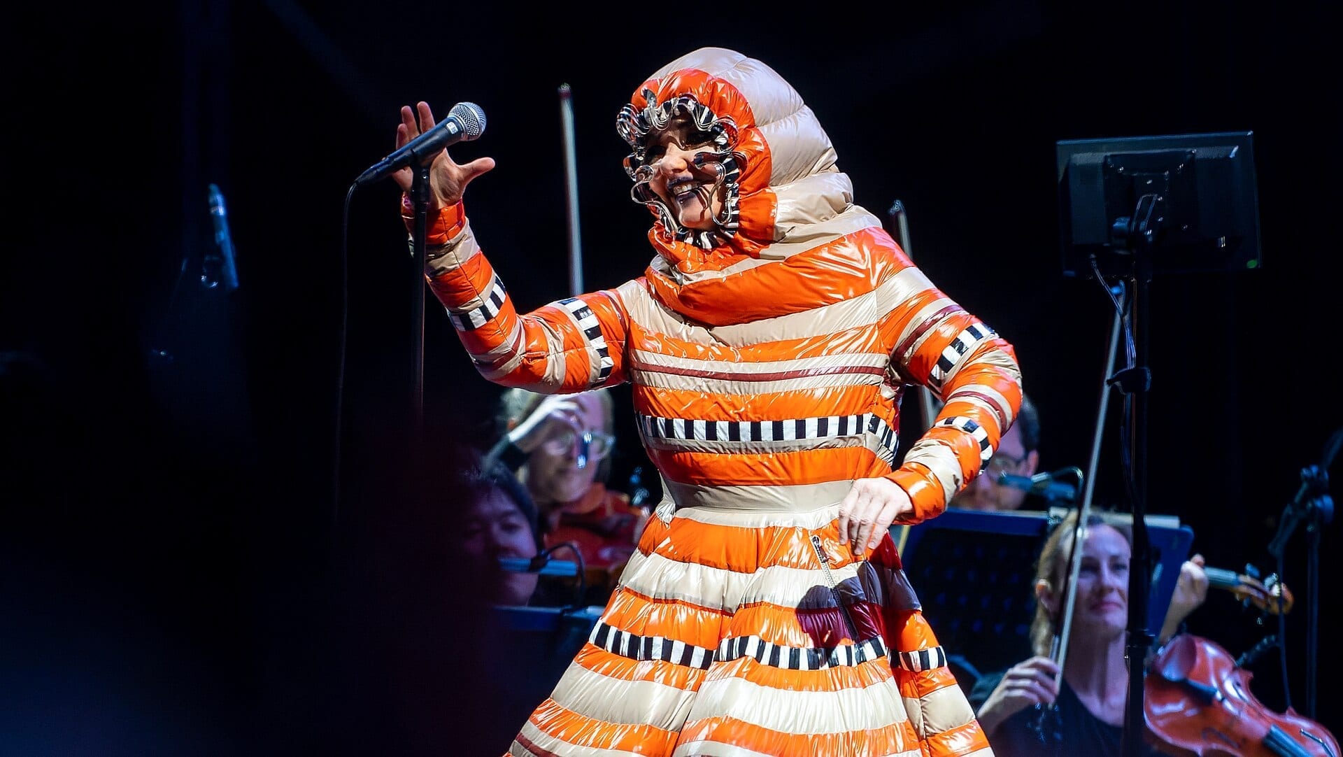 Bjork sings into a microphone in an elaborate pale costume and headpiece, lit against a dark festival stage.