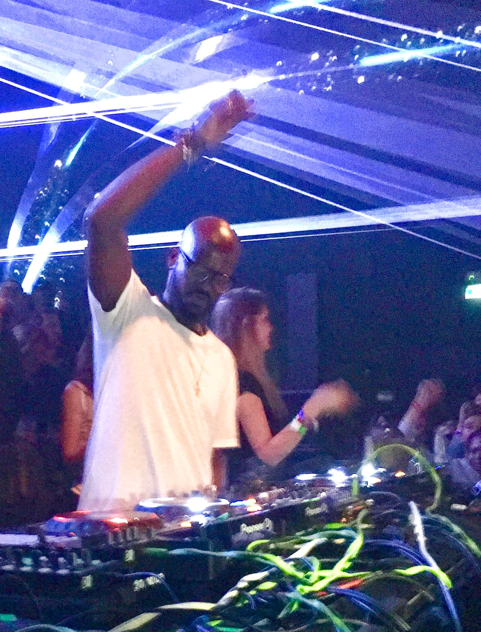 Black Coffee, in a white t-shirt, pumps a fist in the air behind DJ decks under crisscrossing blue laser beams.