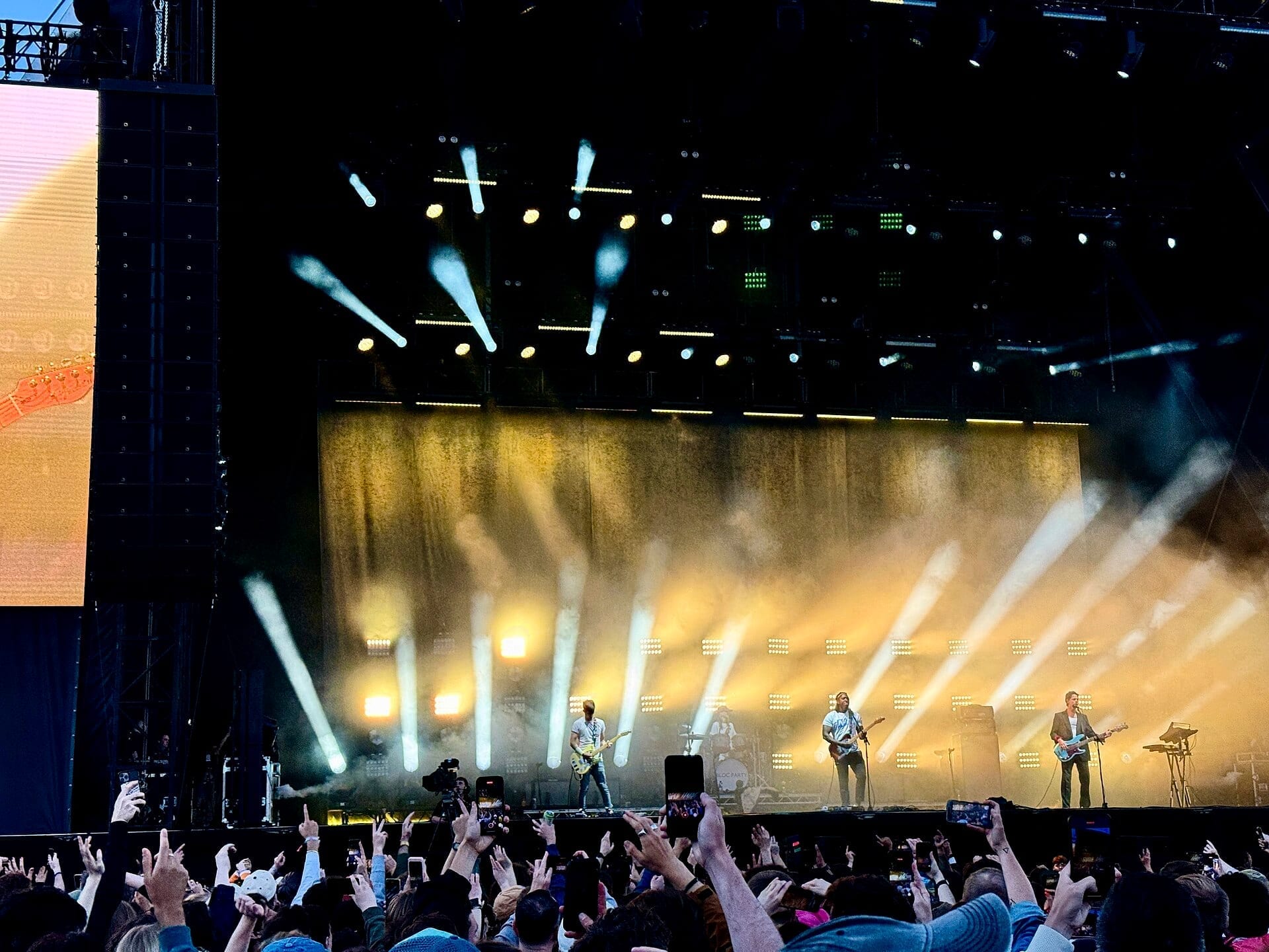 Bloc Party performs on a wide festival stage lit by fanned white spotlights and gold haze, with a crowd holding up phones in the foreground.