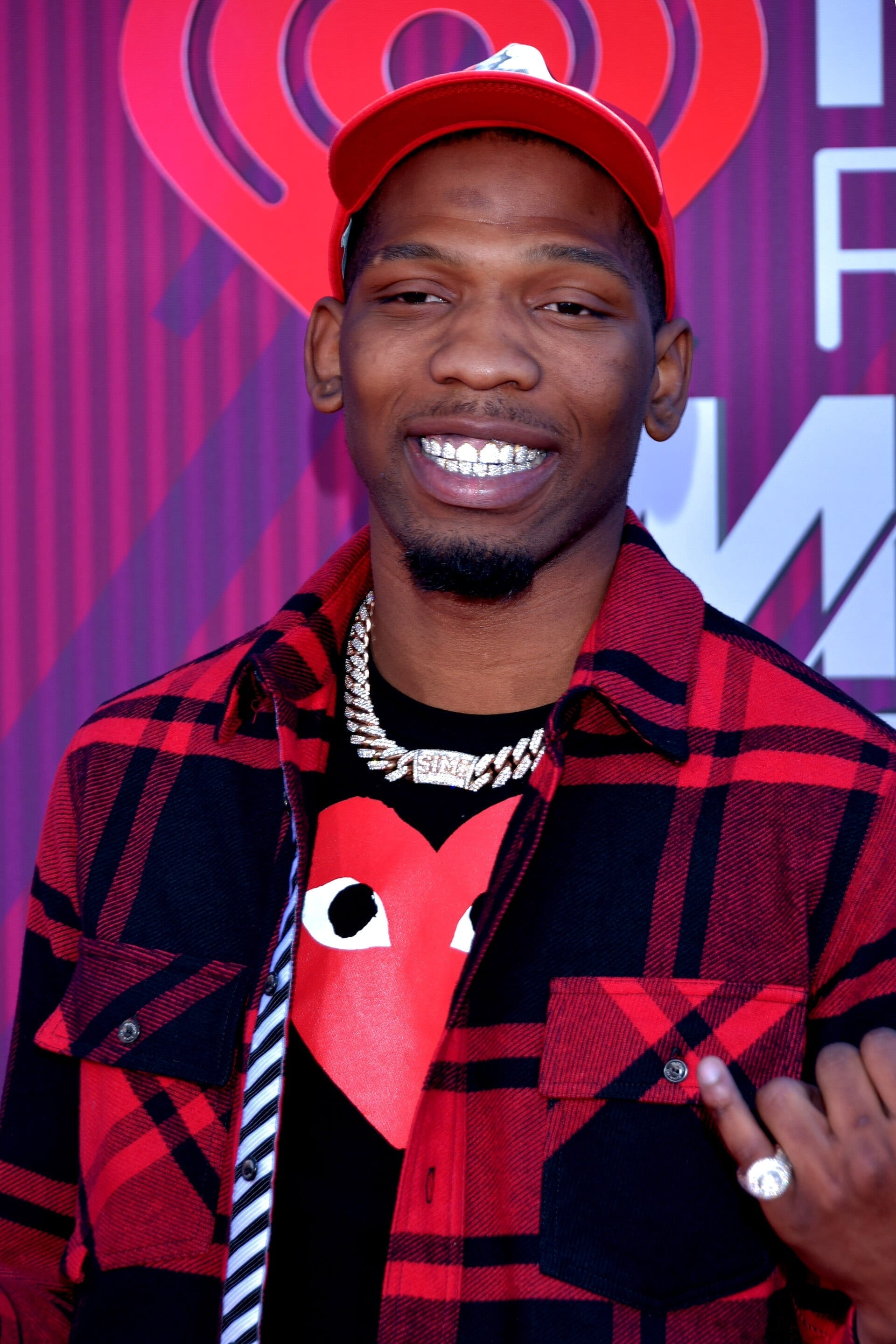 BlocBoy JB smiles for the camera on a red carpet, wearing a red and black plaid jacket over a graphic t-shirt and a red cap, a grill visible in his smile.