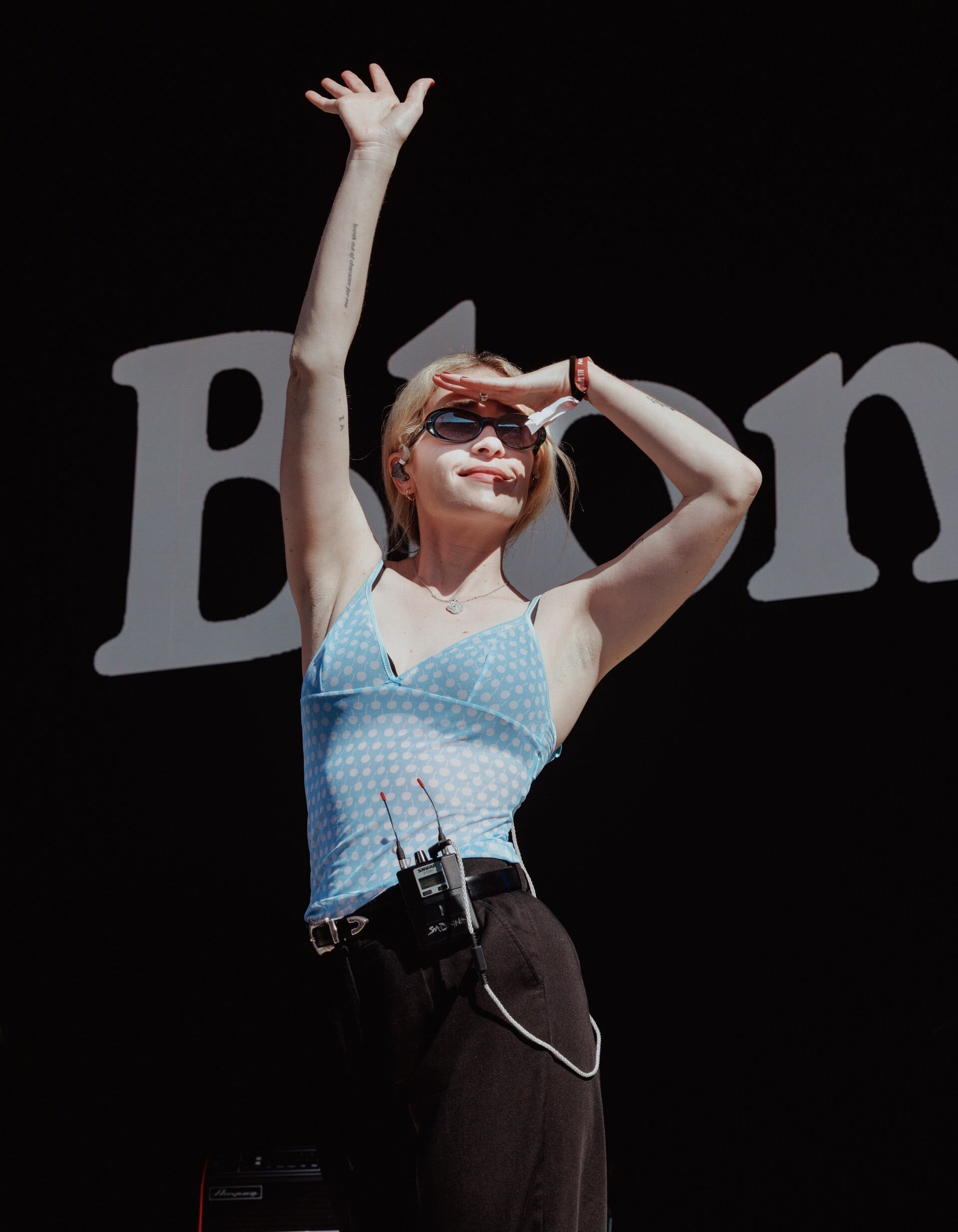 Blondshell, wearing sunglasses and a blue polka-dot top, raises one arm overhead on a sunlit stage in front of a large backdrop with the band's name.