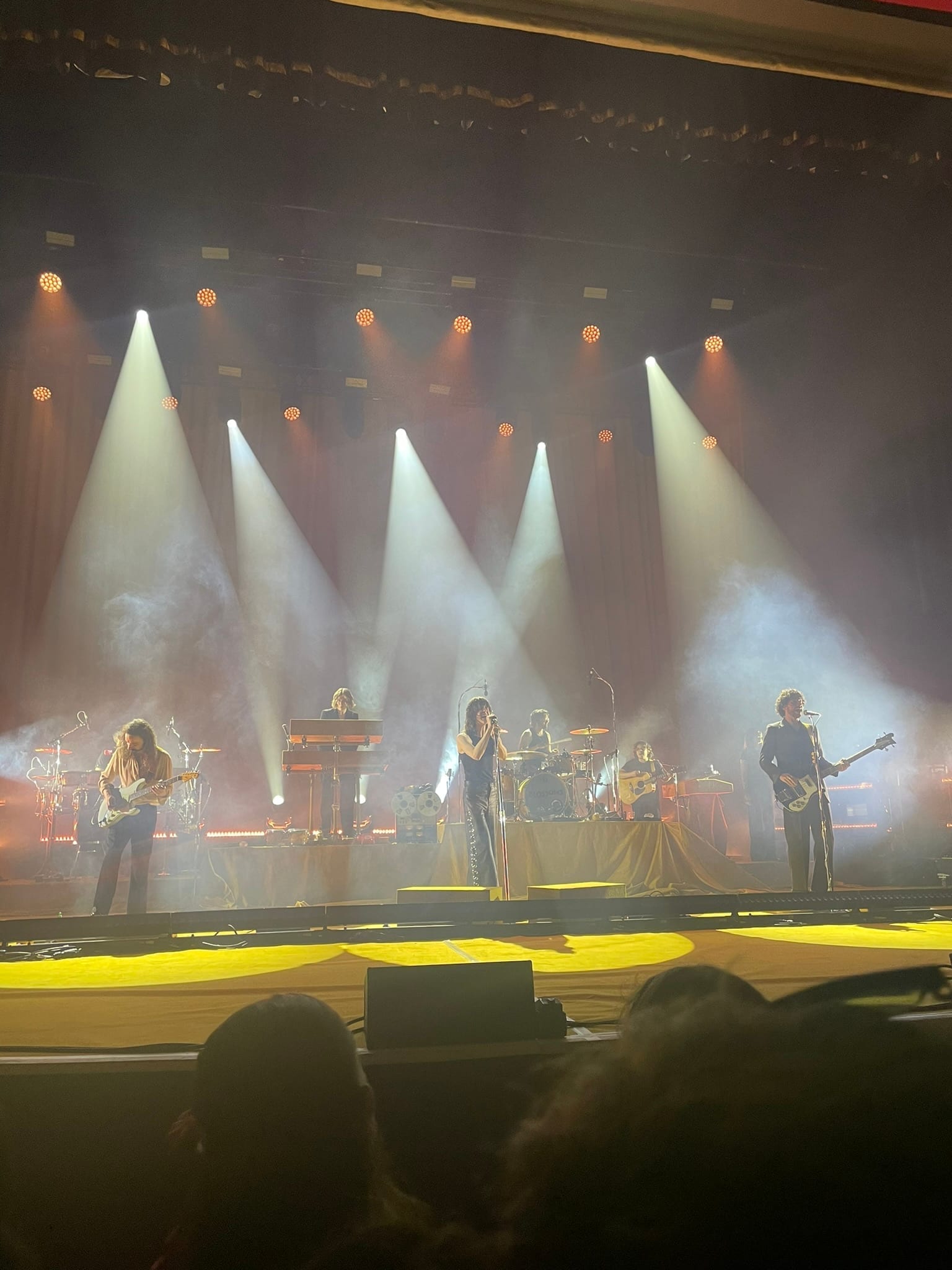 Blossoms perform on a smoky, colorfully lit stage, with band members playing guitar, keyboard, bass, and drums around a singer at the center microphone.