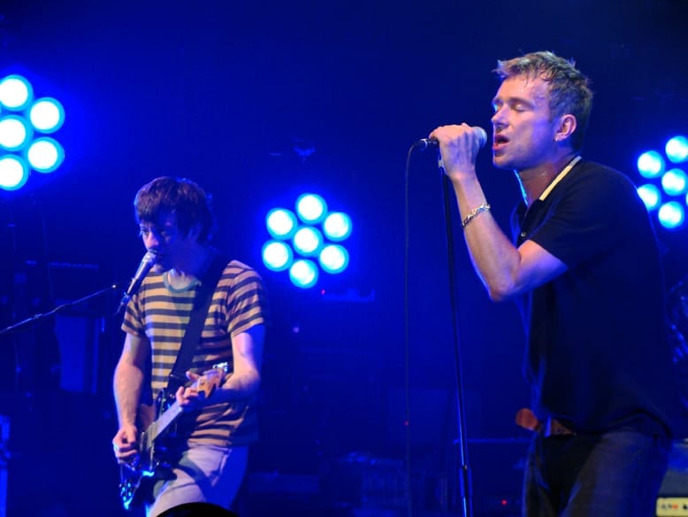 Damon Albarn sings into a microphone stand at right while Graham Coxon plays guitar at left, both under blue stage lights.