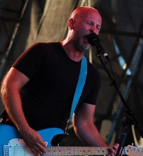 Bob Mould sings with his eyes closed while playing a light blue electric guitar under red stage lighting.