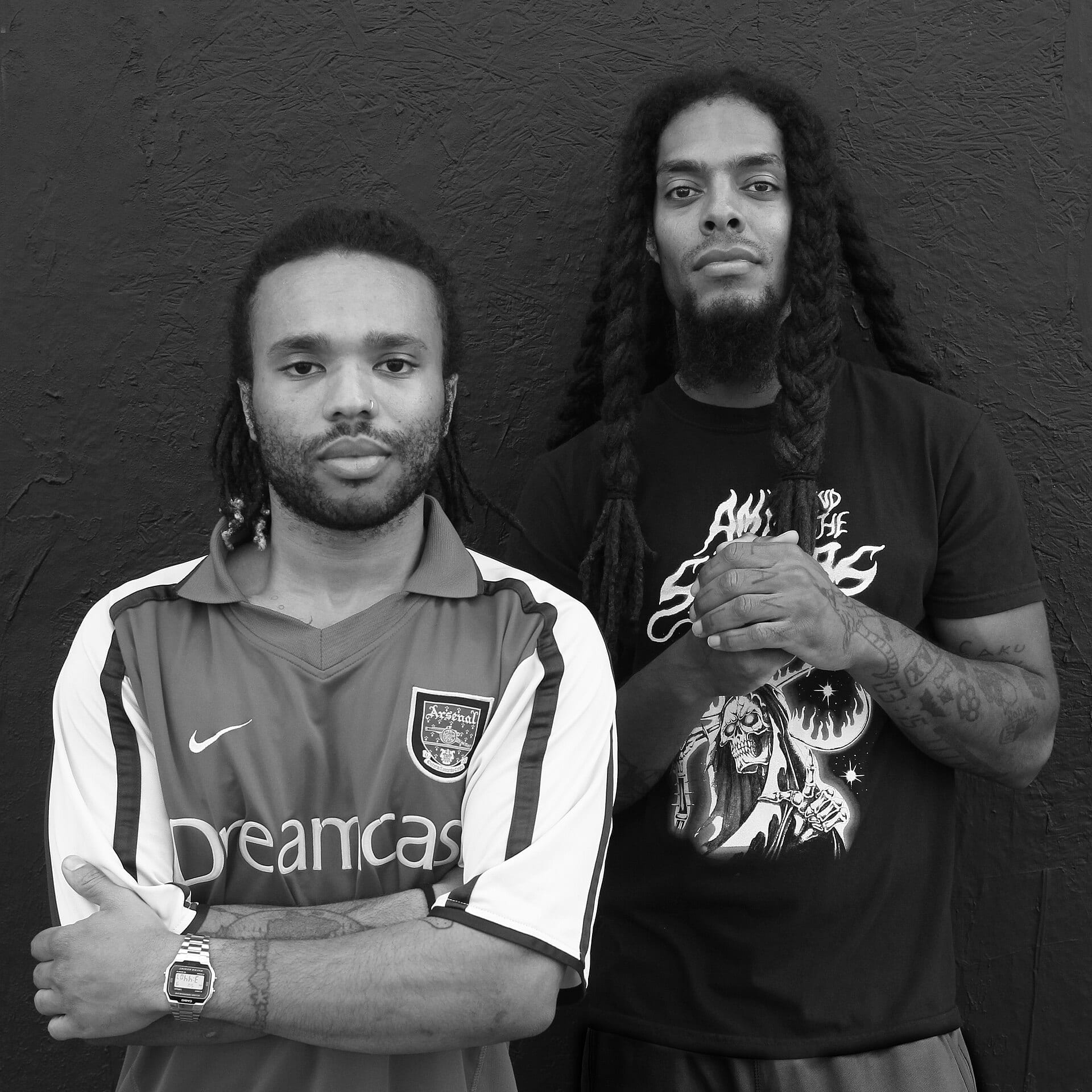 The two members of Bob Vylan pose in black and white against a textured wall, one in an Arsenal Dreamcast jersey with arms crossed, the other in a black graphic t-shirt with clasped hands.