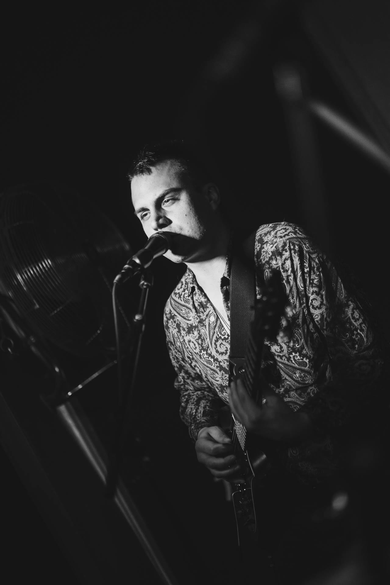 A member of the band Bodega sings into a microphone while playing guitar, photographed in black and white on stage.