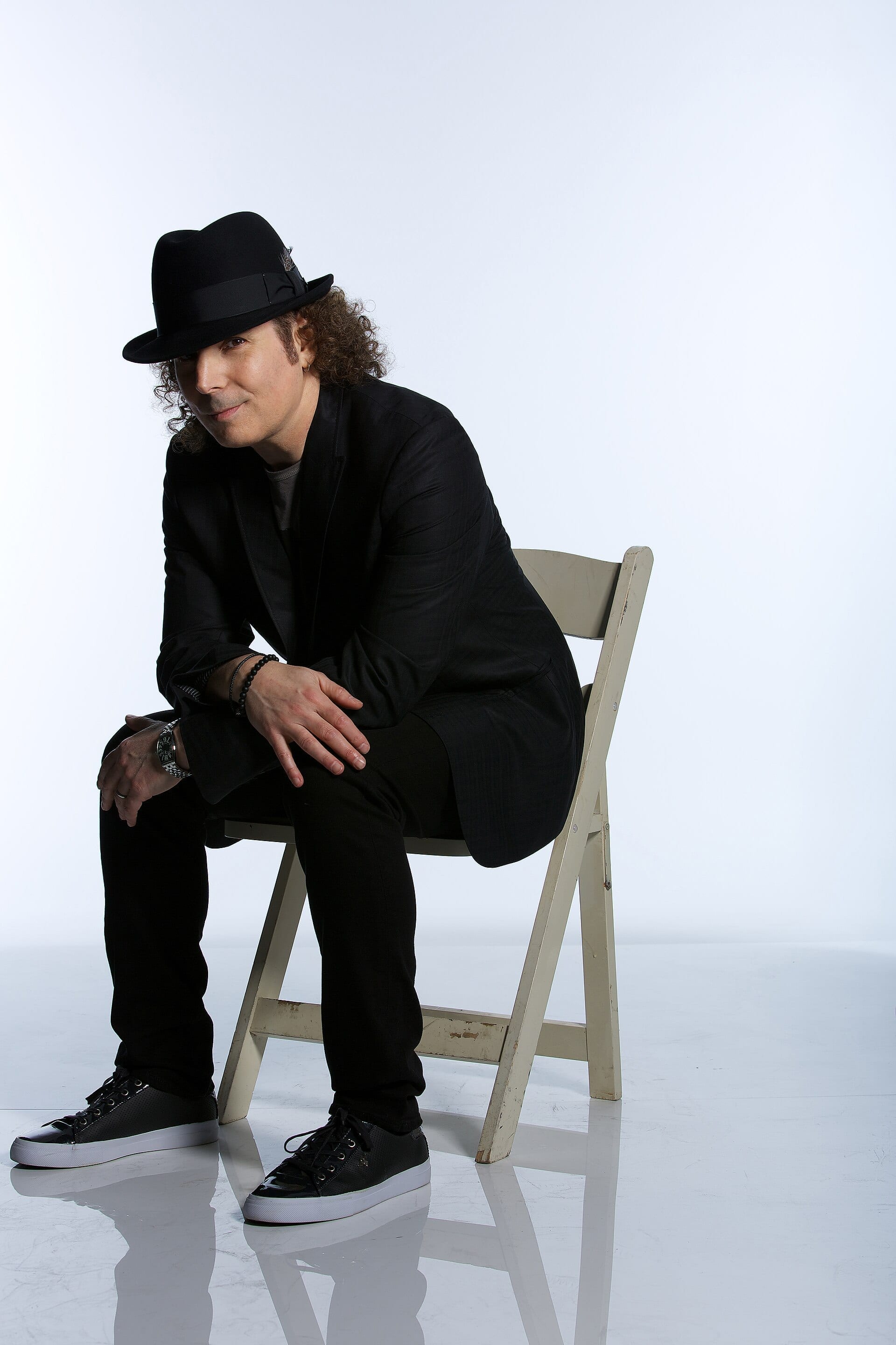 Boney James sits sideways on a white folding chair wearing a black fedora and black suit, resting his clasped hands on his knee.