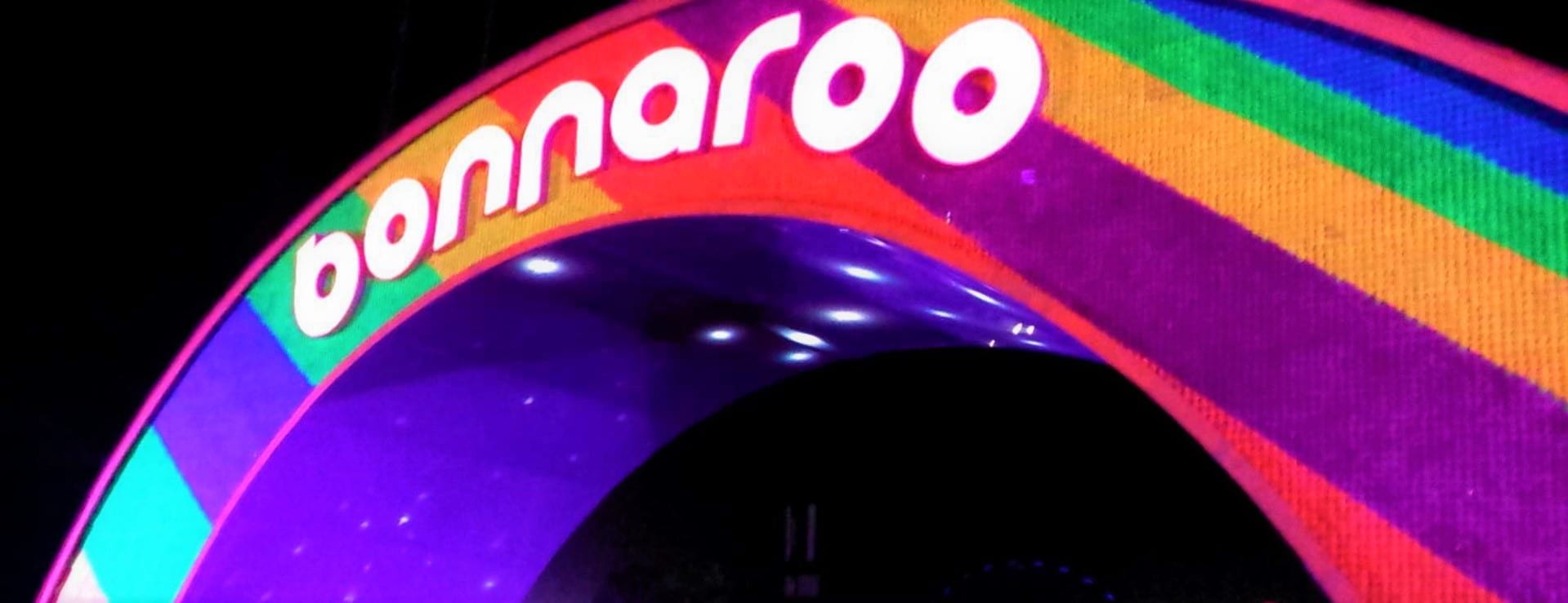 The Bonnaroo arch lit up at night in rainbow stripes, the festival name across it in white lowercase letters.