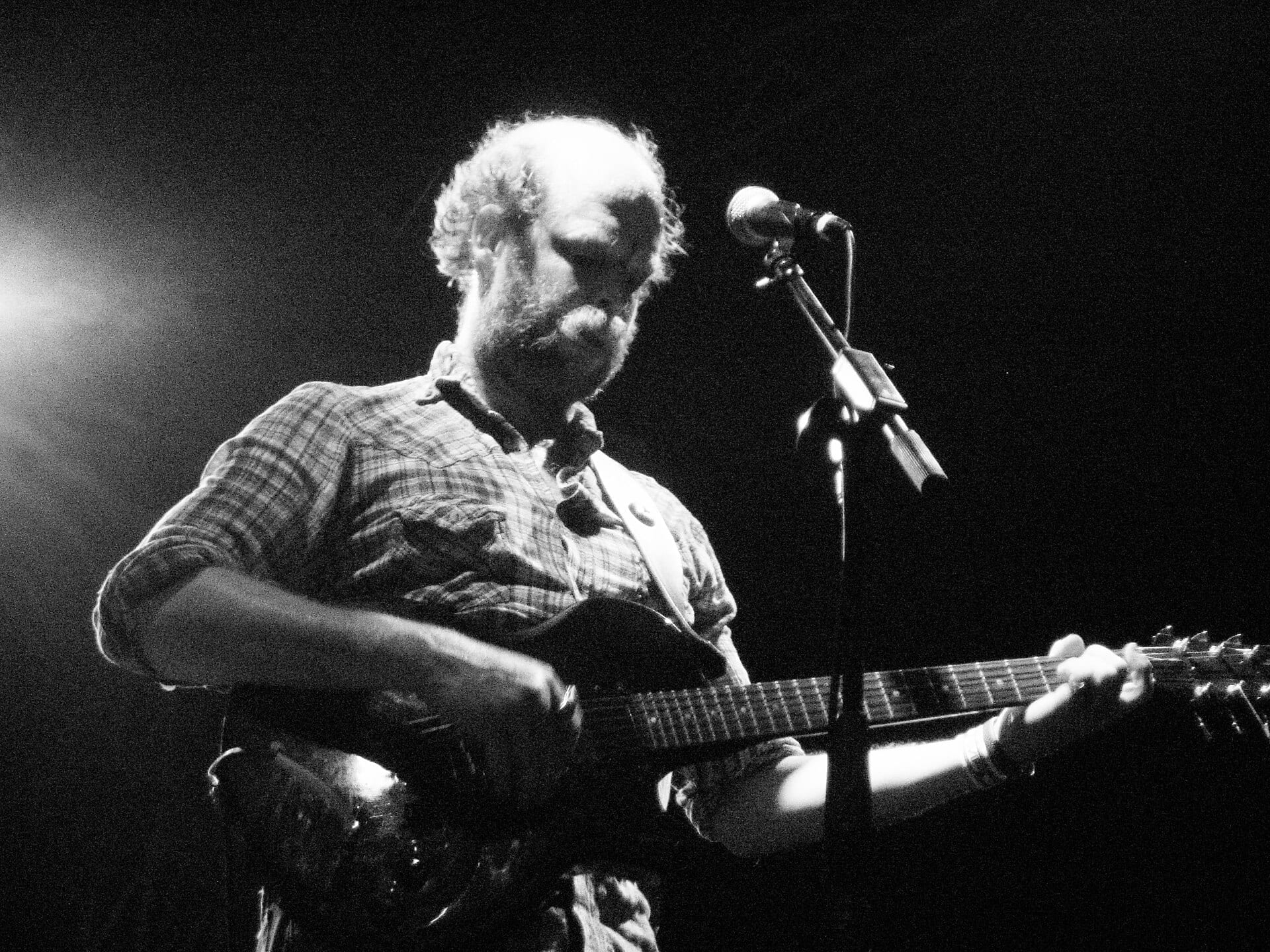 A black-and-white photo of a bearded man playing an electric guitar and leaning into a microphone stand on a dark stage.