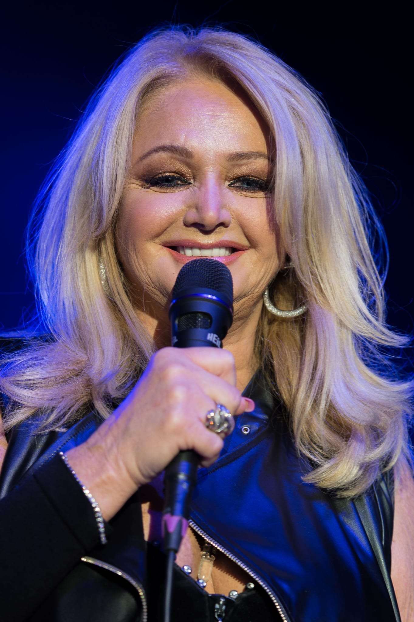 Bonnie Tyler, smiling with long blonde hair and a black leather outfit, sings into a microphone under blue stage lighting.